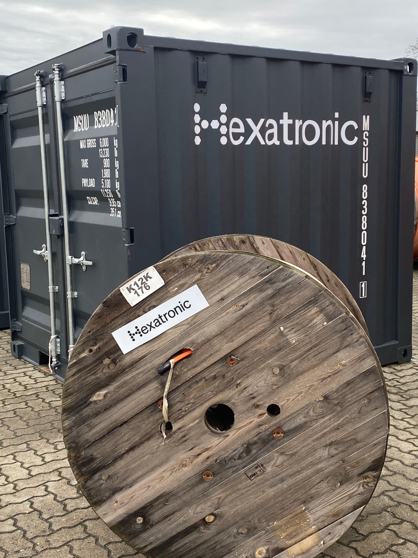 Schwarzer Frachtcontainer mit Hexatronic Aufschrift. Eine Kabeltrommel aus Holz steht davor. 