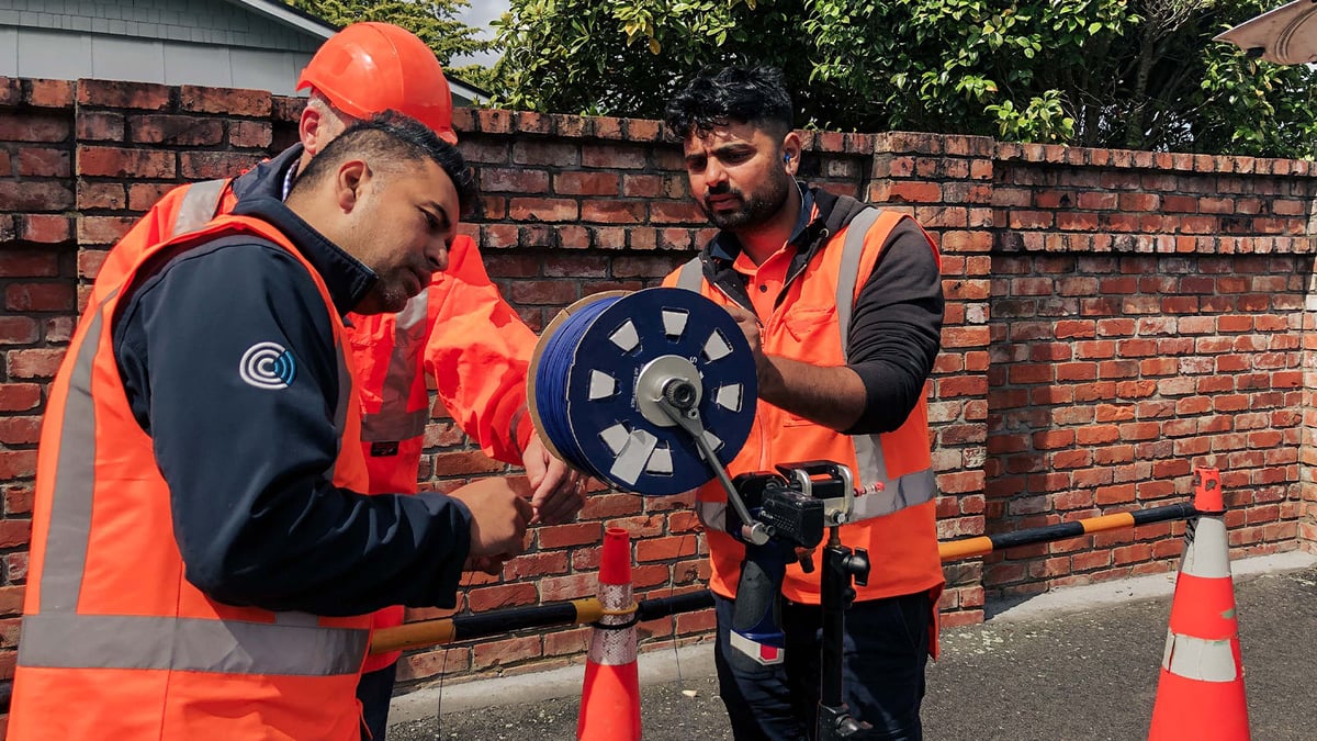 Three people install Stingray Air Blown Fiber. In the background, a house and green trees can be seen behind a red brick wall.