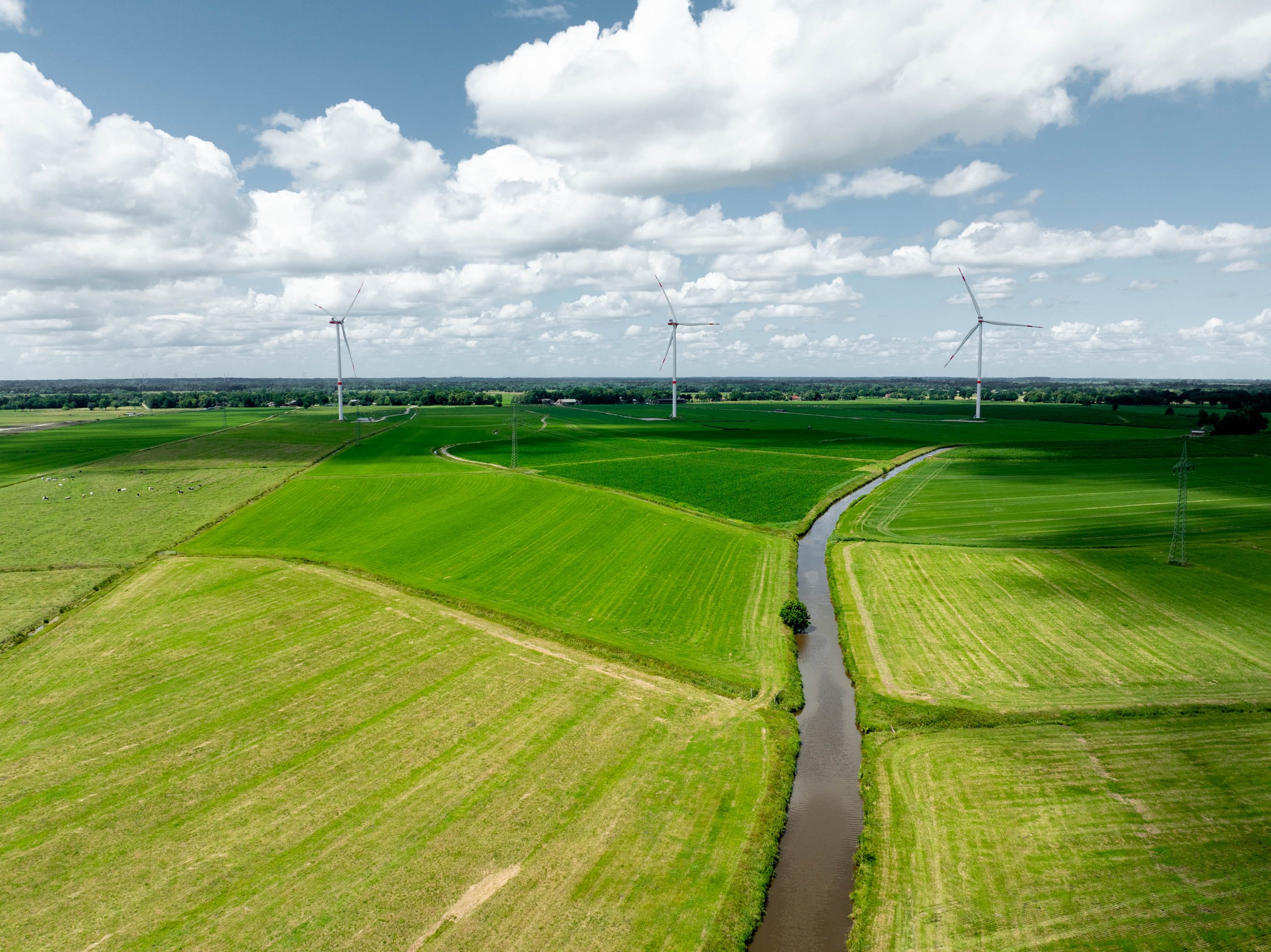 Green fields with a winding river and three wind turbines under a partly cloudy sky.
