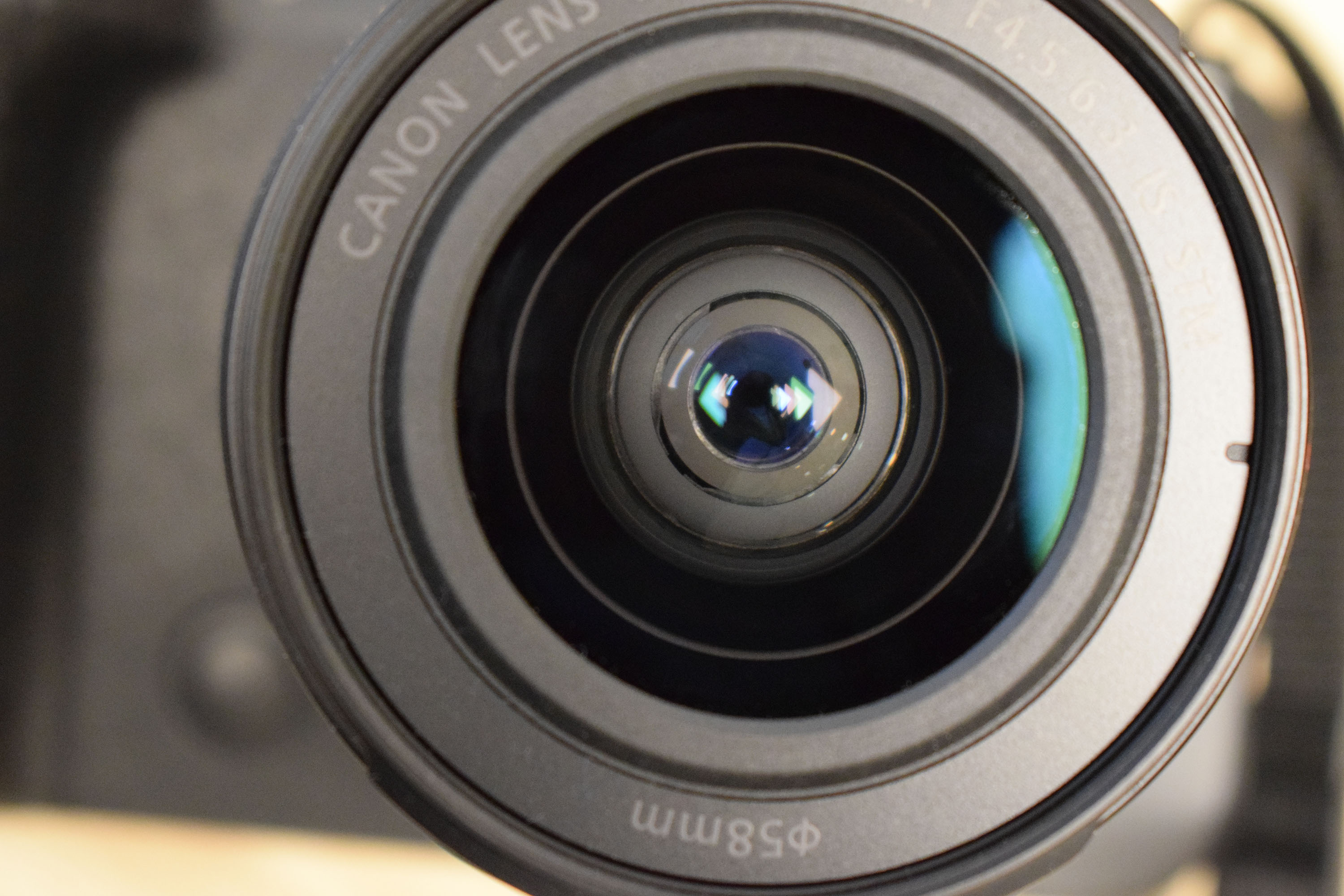 Close-up of a Canon camera lens, showing intricate details and light reflection on its glass surface. Background is indoors, softly lit.