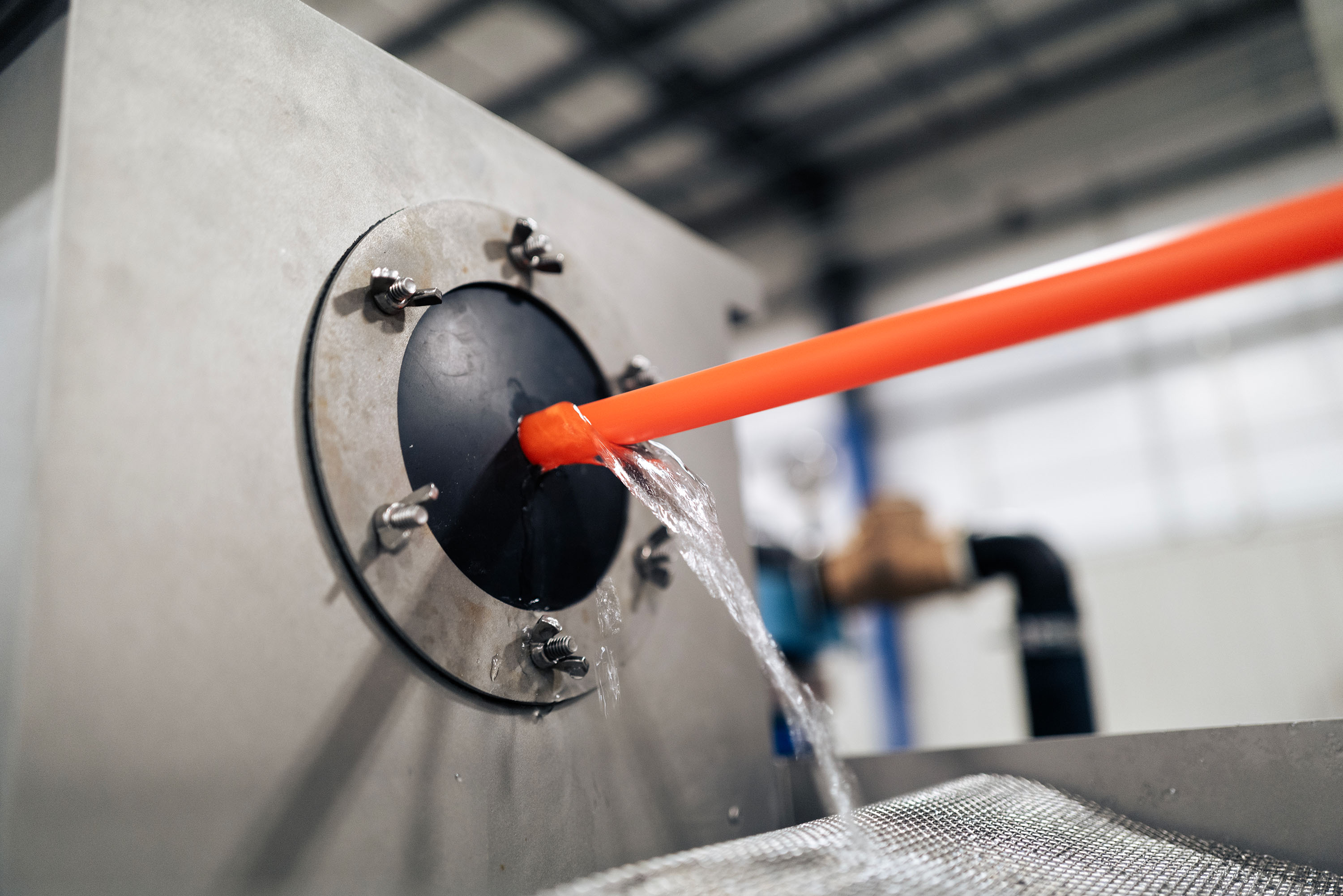 A close-up of an industrial machine that produces a micro duct. The machine has a metallic exterior and water flows around the orange duct.