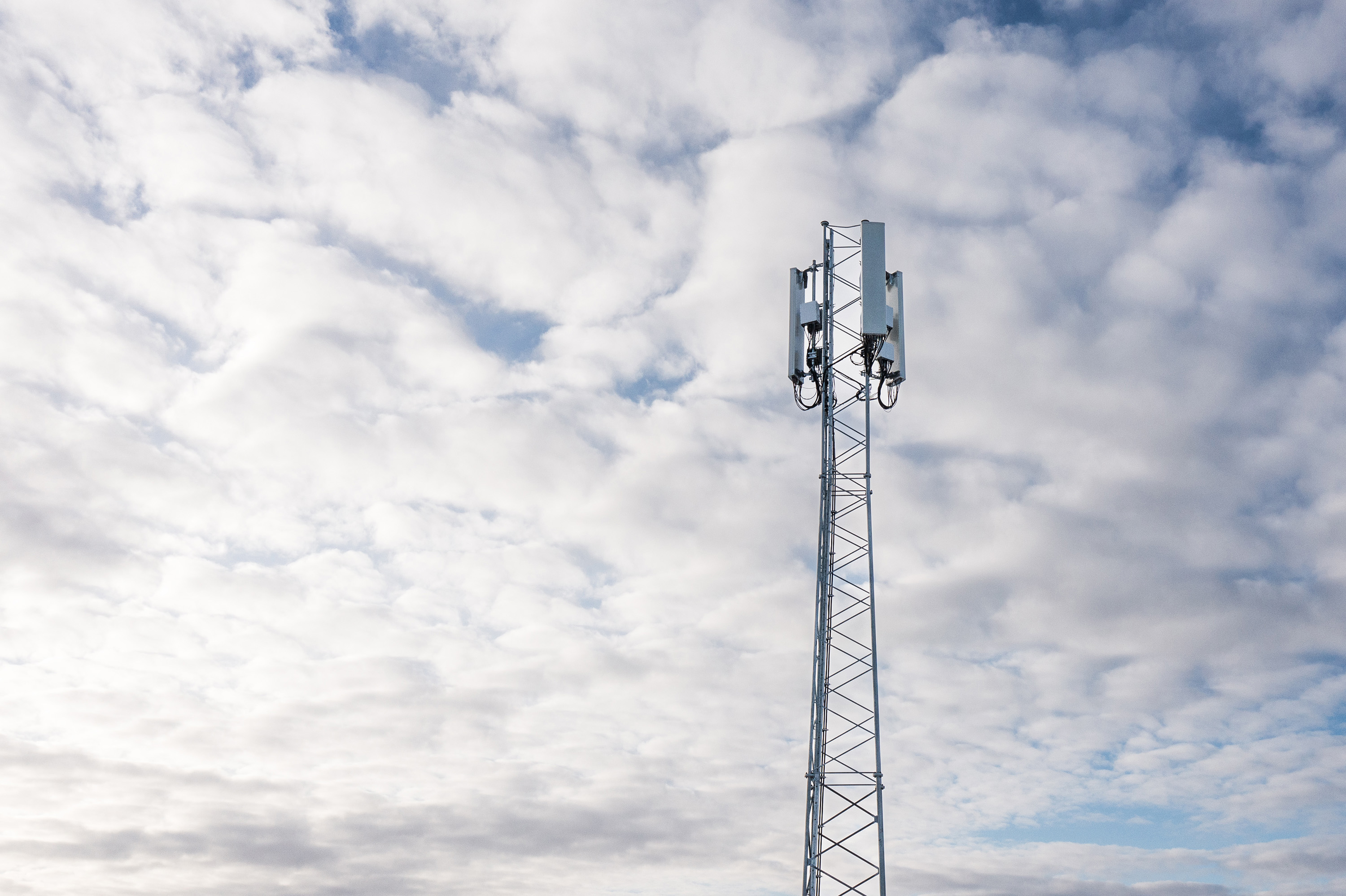 A tall metal tower with 5G antennas and communication equipment mounted on top. The sky in the background is filled with white clouds.