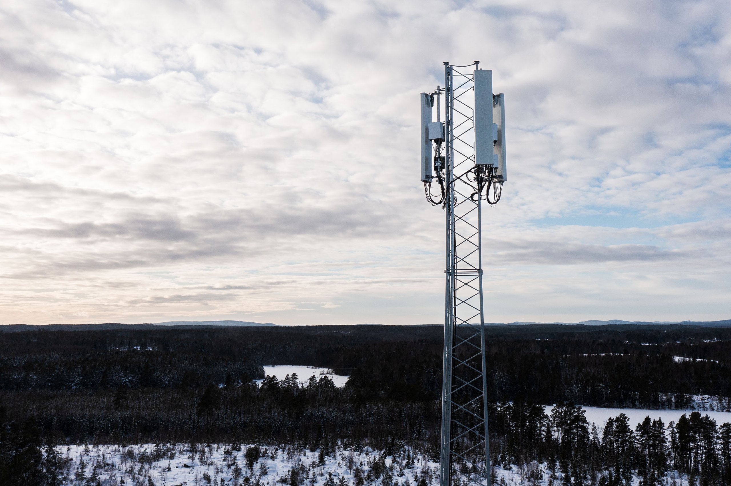 A cell tower in a snowy forest. The tower is equipped with 5G antennas and equipment. The forest is barren and covered in snow.