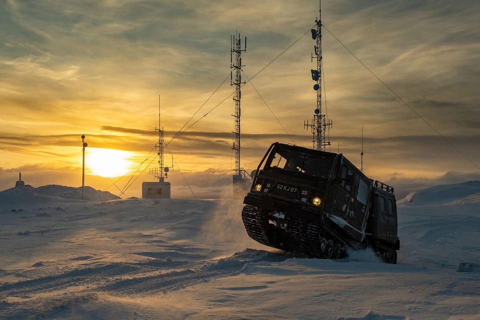 A tracked vehicle moves across snowy terrain at sunset, near communication towers, evoking a sense of remote, harsh yet beautiful environment.
