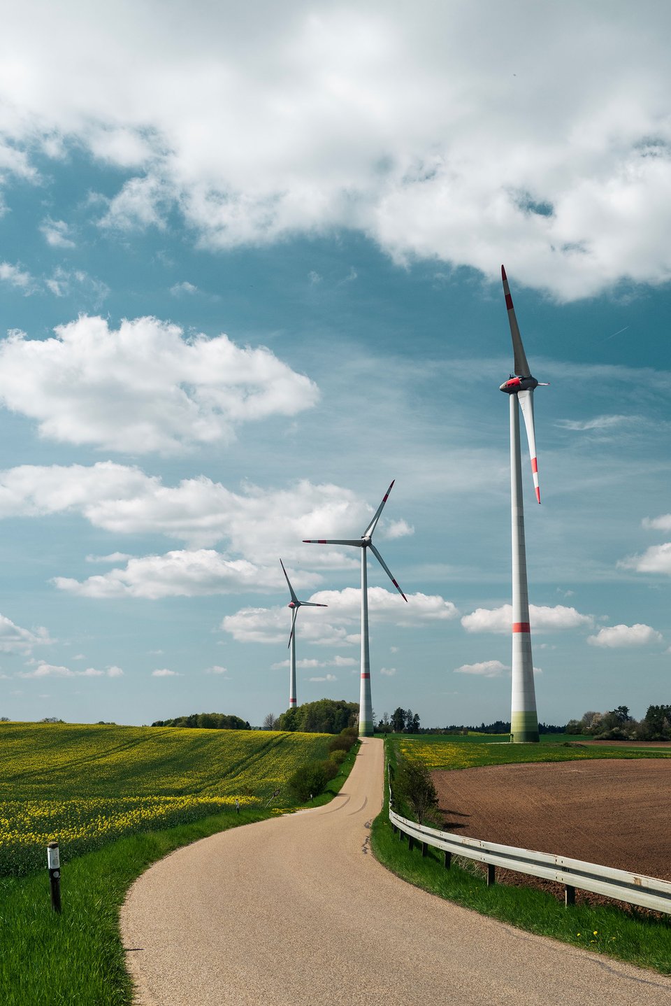 A rural area with three wind turbines standing tall on a green field next to a country road. The partly cloudy sky adds depth to the scene.
