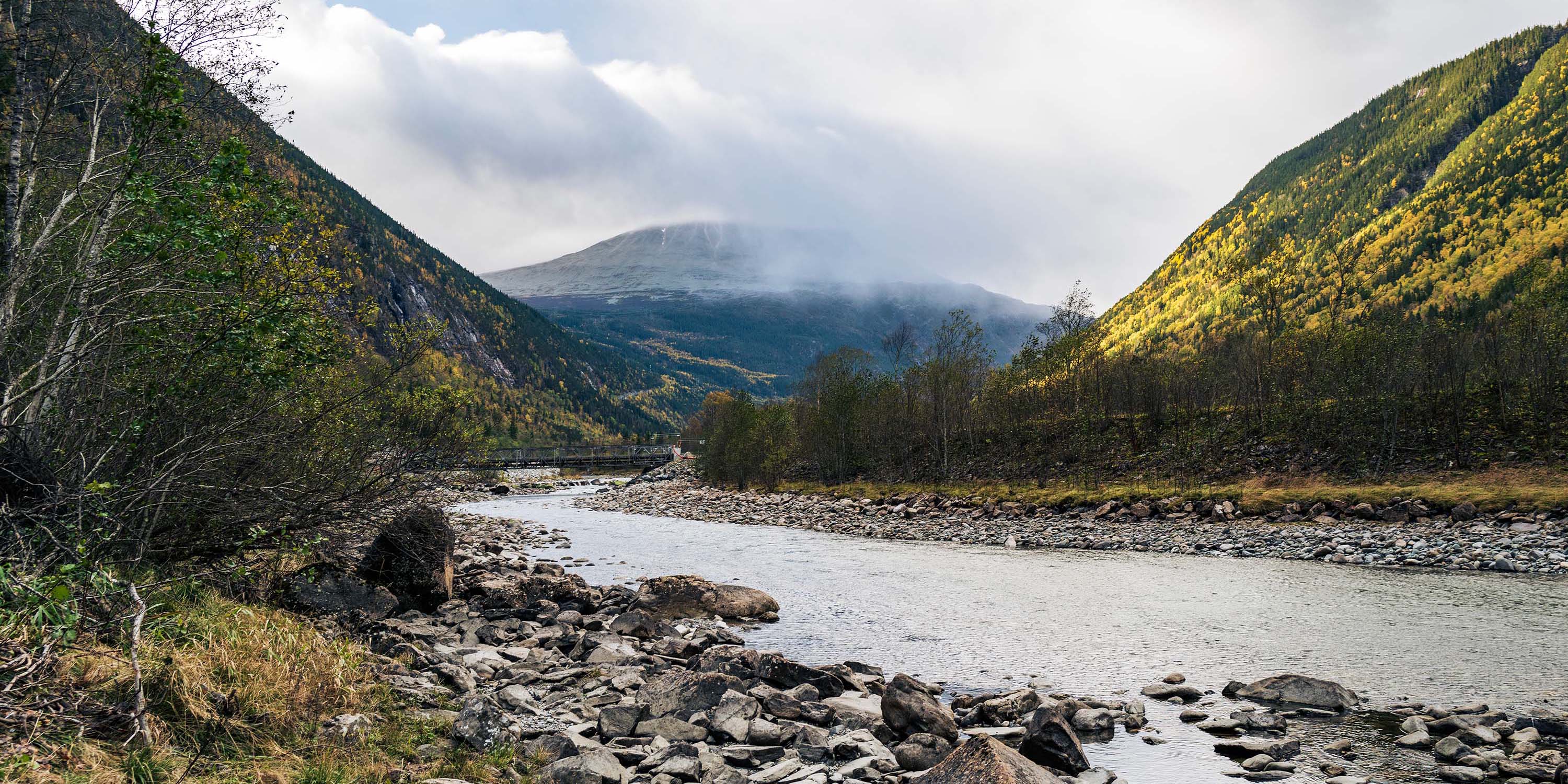 A serene river flowing through rocky mountains under a partly cloudy sky. The water is clear and the banks are rocky.