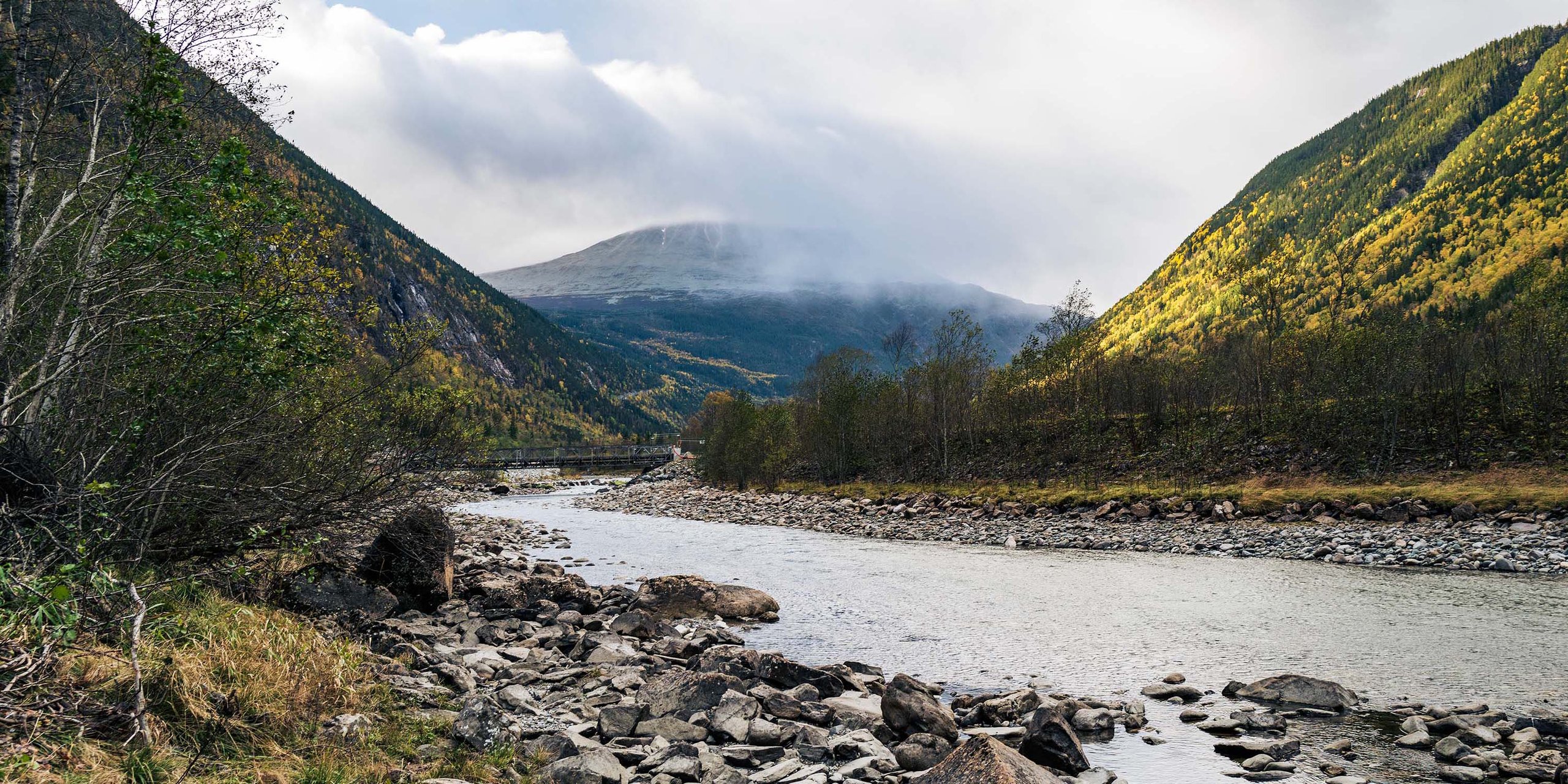 A serene river flowing through rocky mountains under a partly cloudy sky. The water is clear and the banks are rocky.