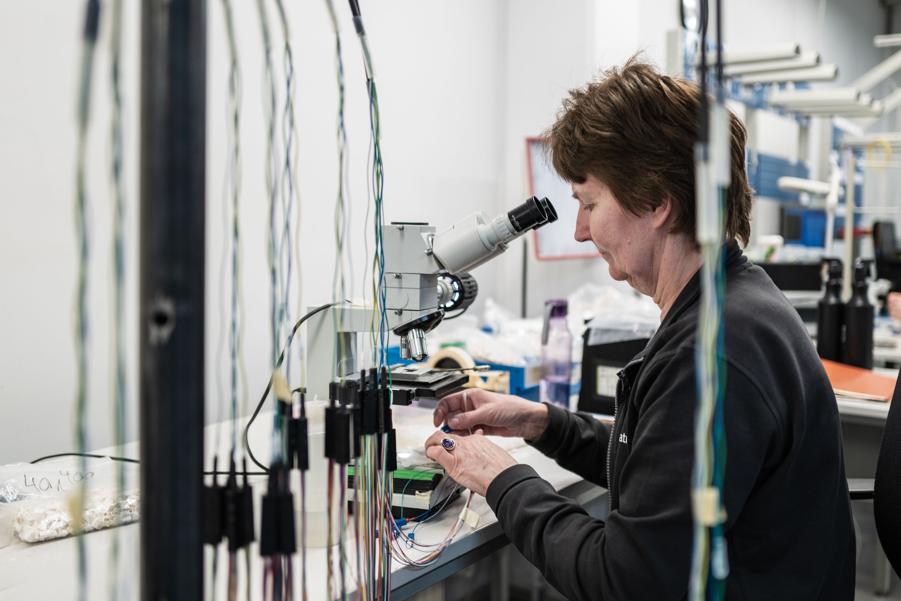A person working on intricate wiring using a microscope in a well-equipped lab. Various colored wires and fiber optic components are visible.