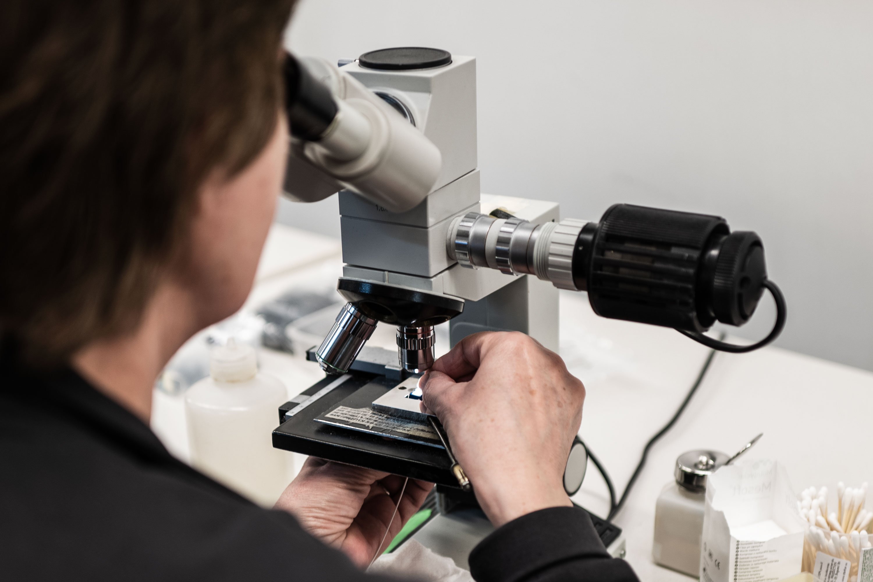 A person uses a microscope to take a closer look at a fiber optic wire, in a well-lit laboratory There are containers and cotton swabs to the right.