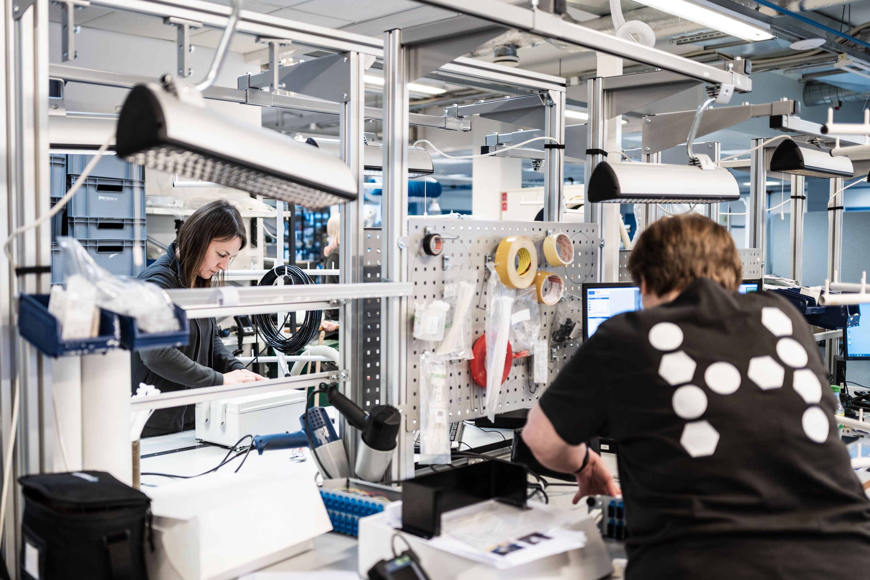 Two people are working at separate stations, one appears to be inspecting or assembling something while the other is focused on a computer screen.