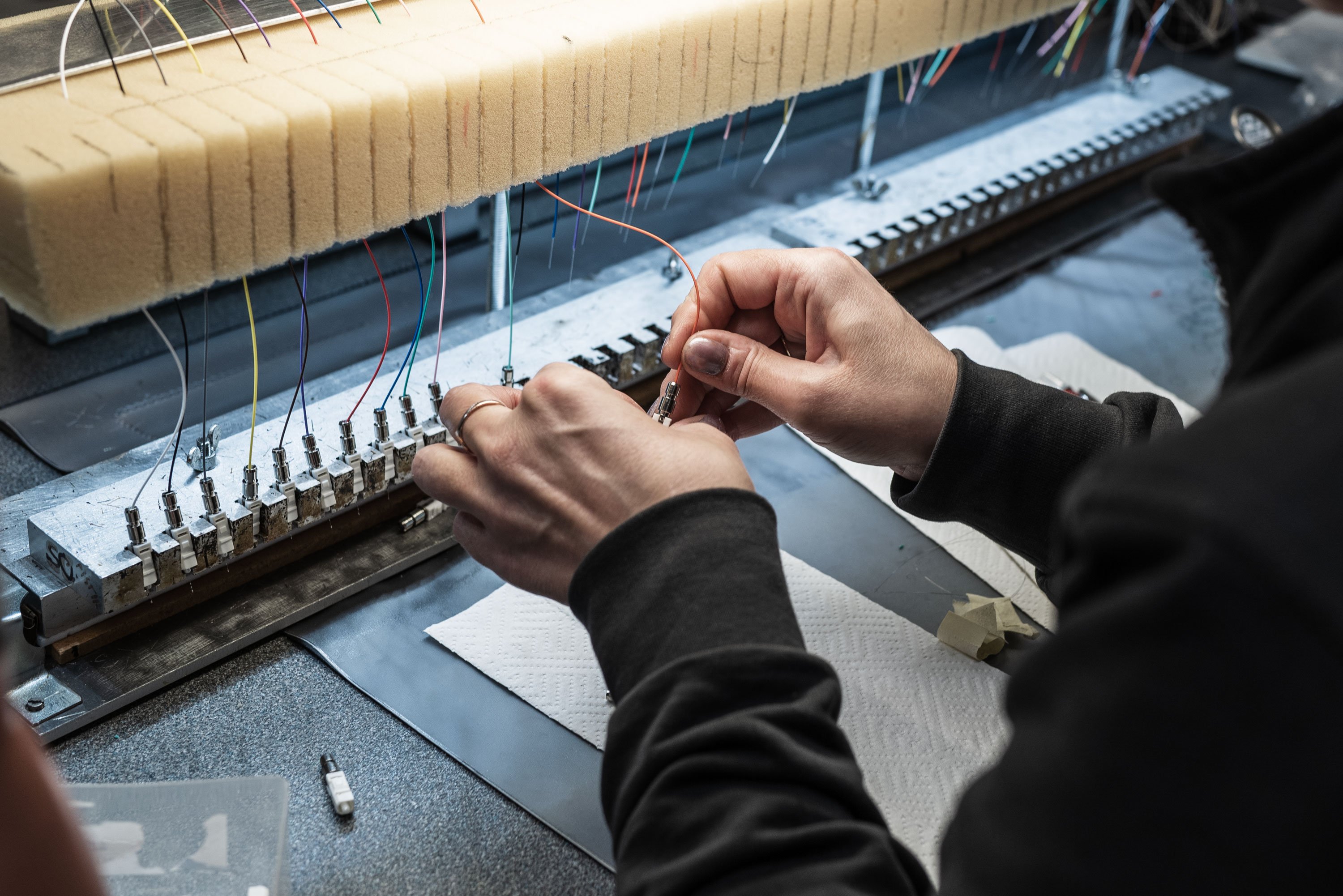 This image shows a person’s hands working on a complex array of colorful fiber optic wires connected to a machine in an industrial setting.