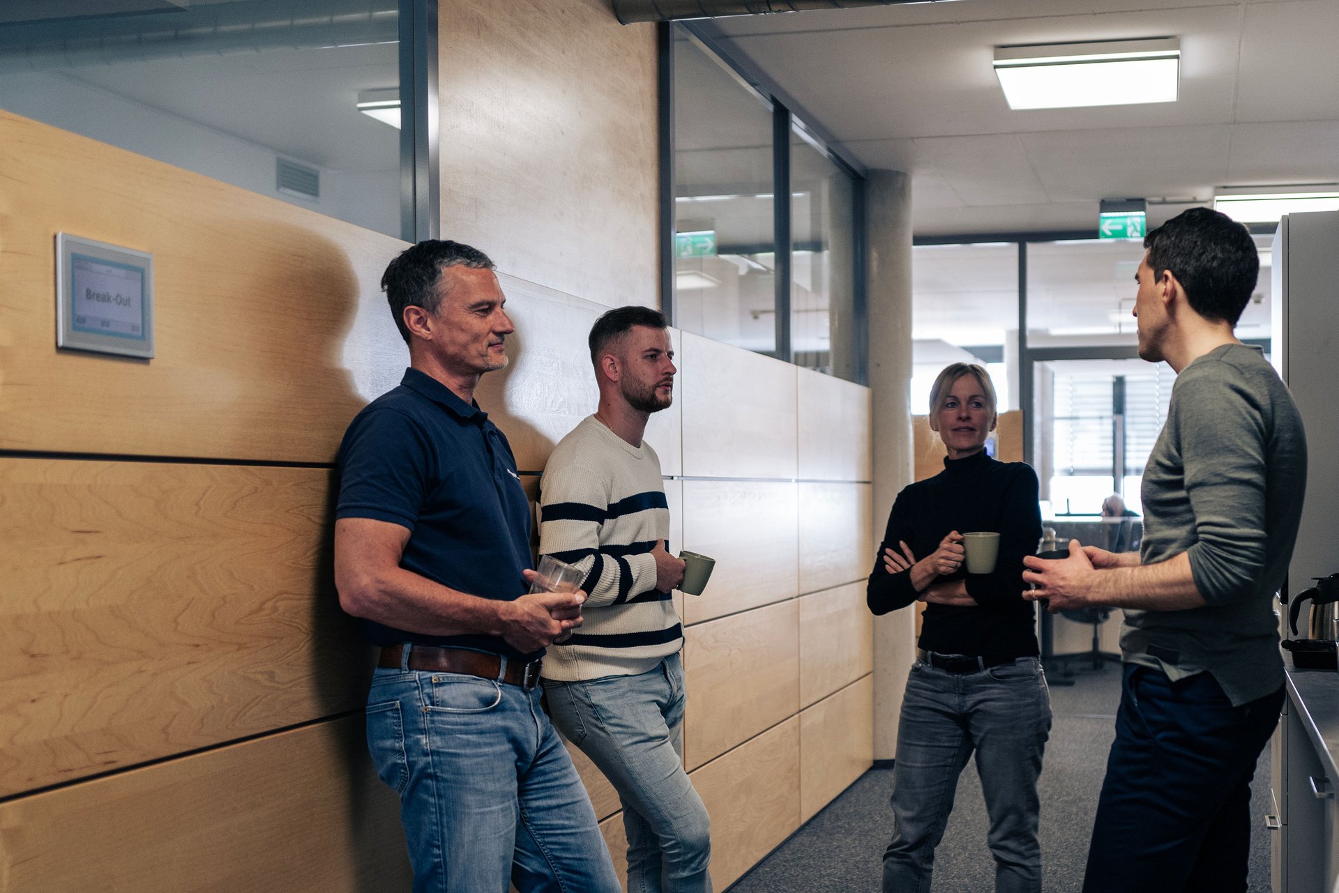Four people are standing and talking in a modern office hallway, holding coffee mugs. It is wooden panels on the walls and ceiling pipes visible.