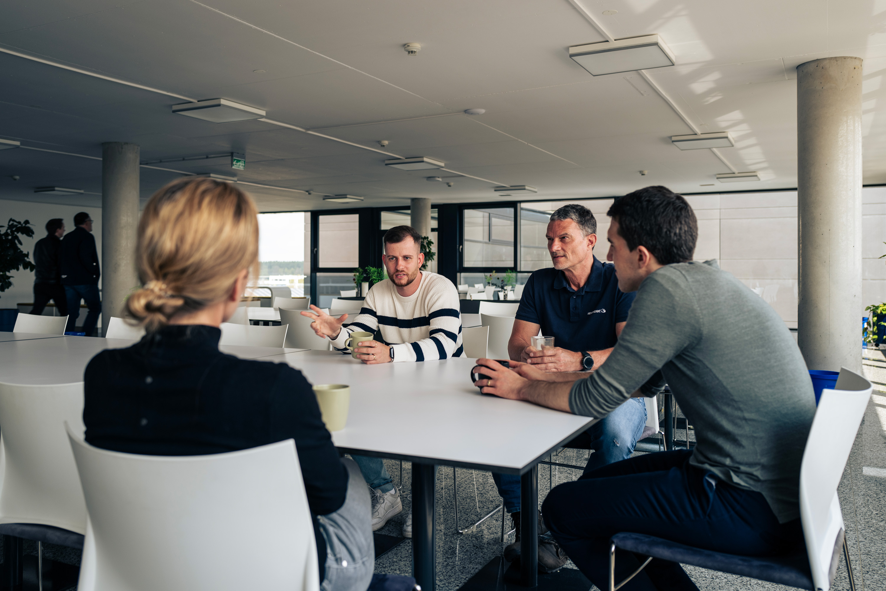 The image shows four people sitting around a white table in a bright, modern room with large windows and plants, engaging in a discussion.