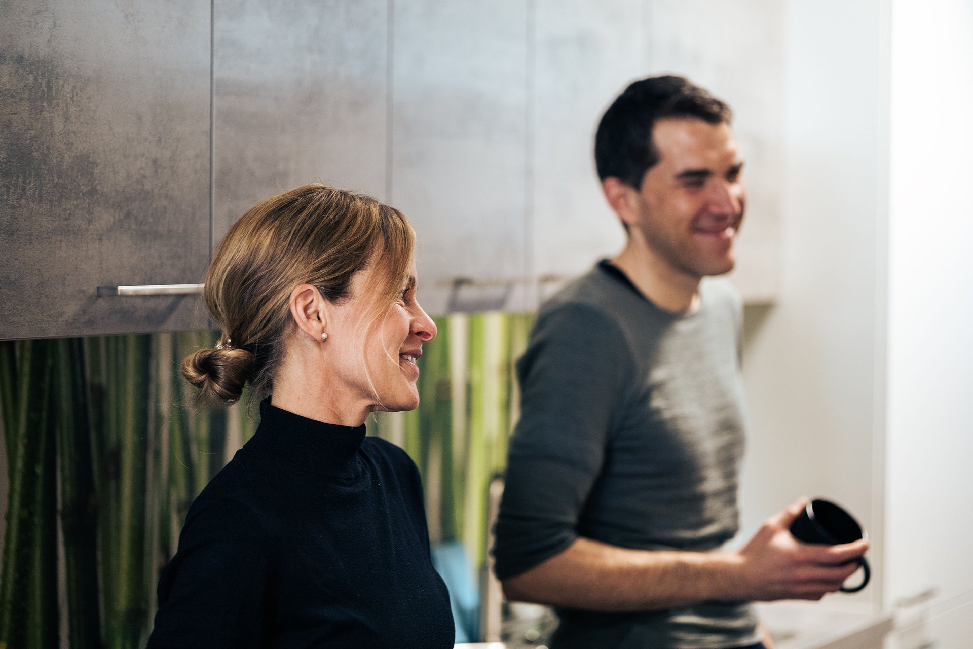 Two people stand next to each other in a coffee room and appear to be talking to someone opposite. One has a black coffee cup in his hand.