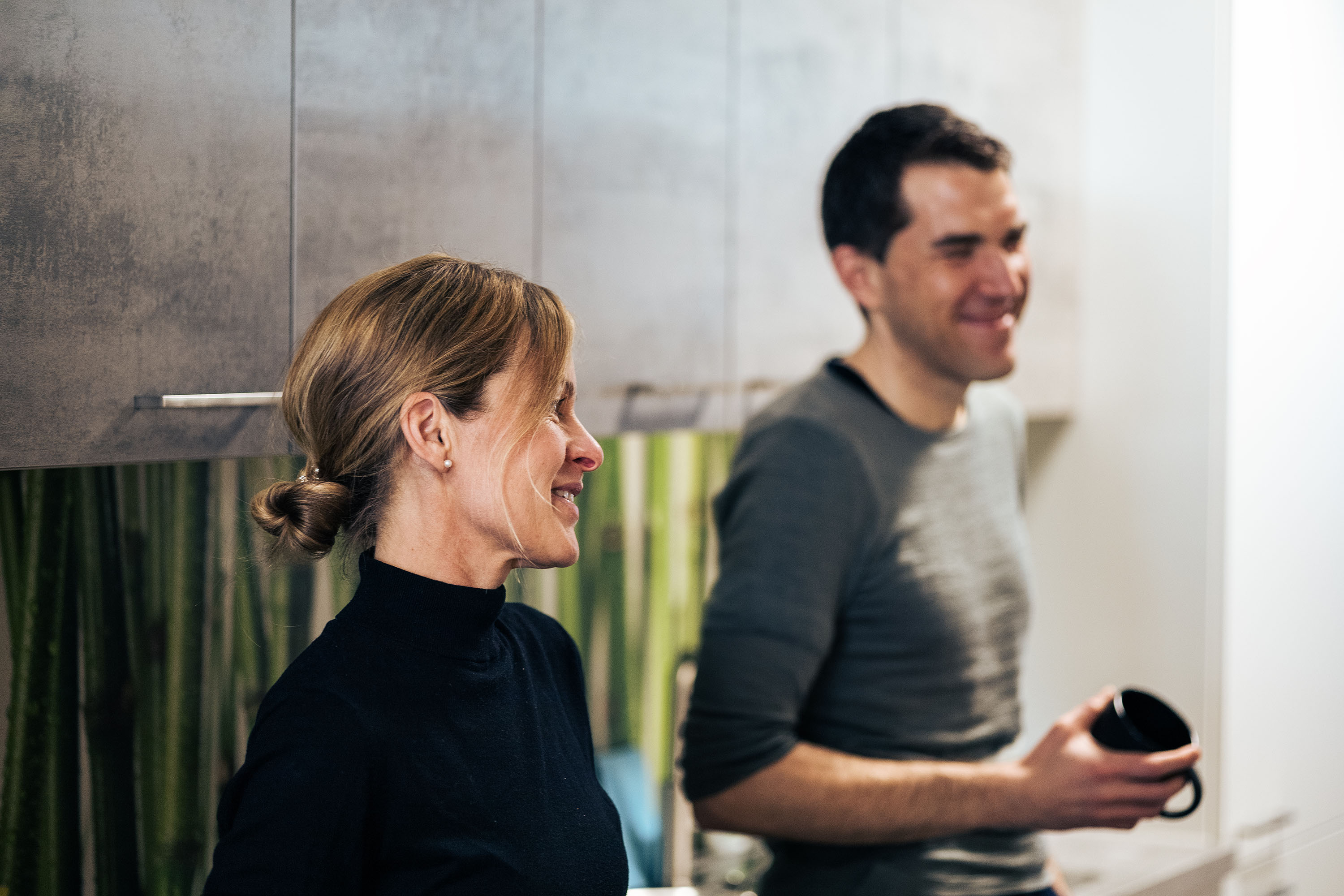 Two people stand next to each other in a coffee room and appear to be talking to someone opposite. One has a black coffee cup in his hand.