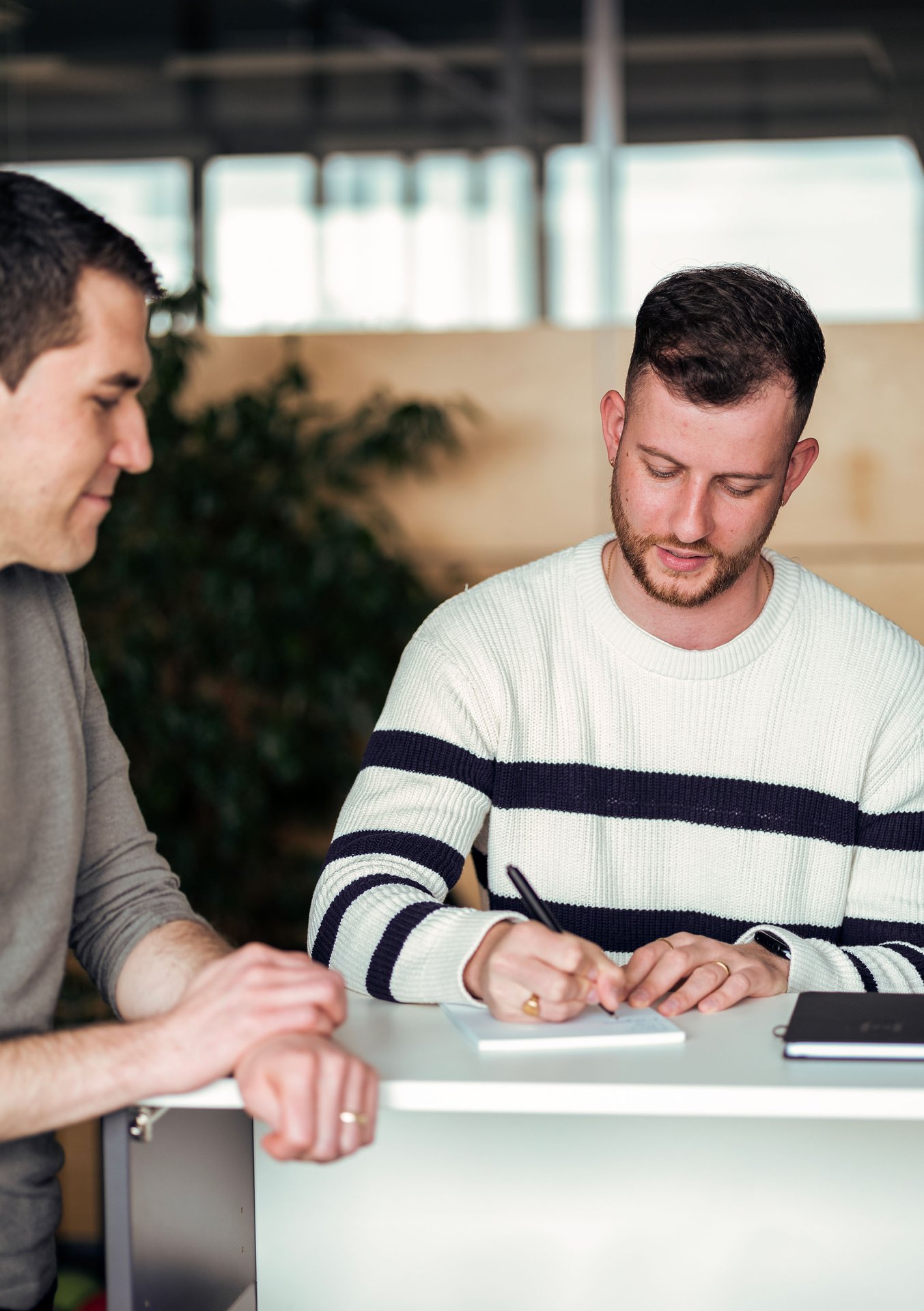 Two people are indoors. One is writing in a notebook, the other is observing. They are standing near a white table.
