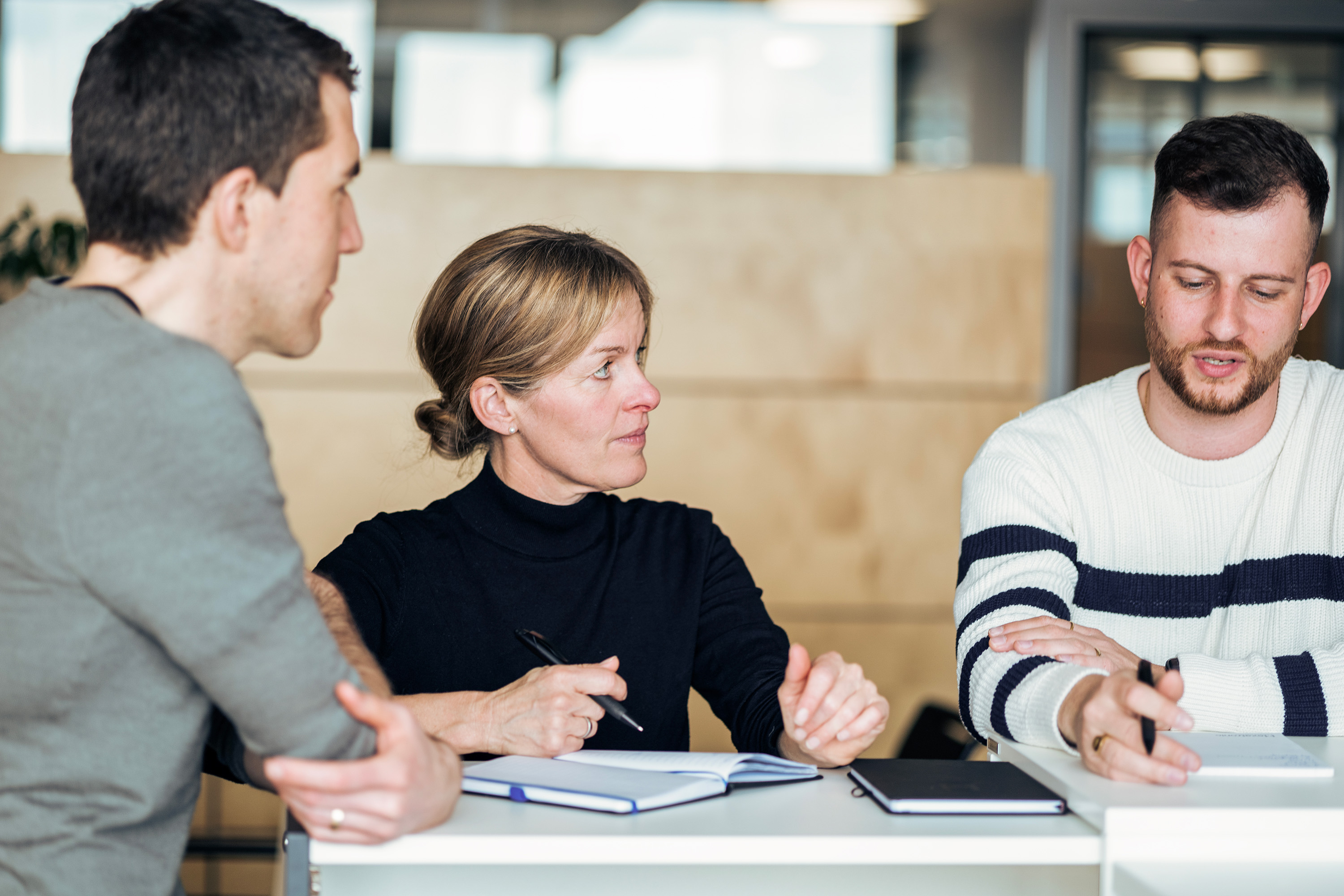 Three people are discussing something around a table with notebooks in a well-lit indoor setting.