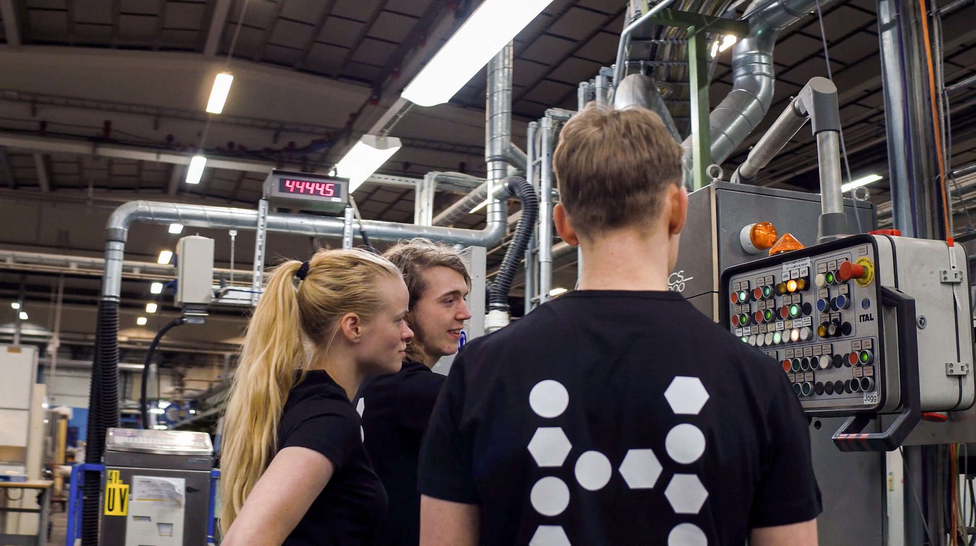 Three people in black attire are observing a complex machine with numerous buttons and switches in an industrial setting.