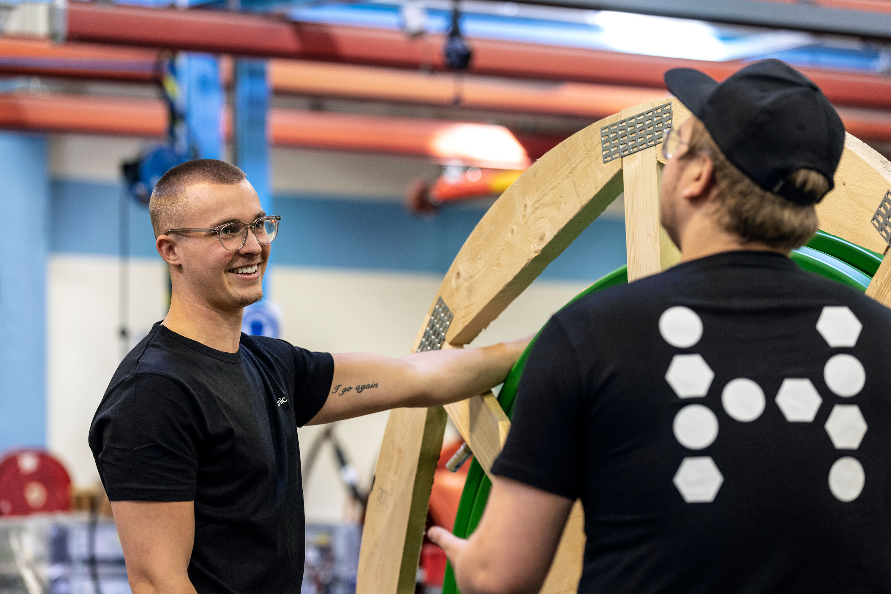 Two people are standing in an industrial room next to a large wooden drum wrapped with microduct and talking to each other.