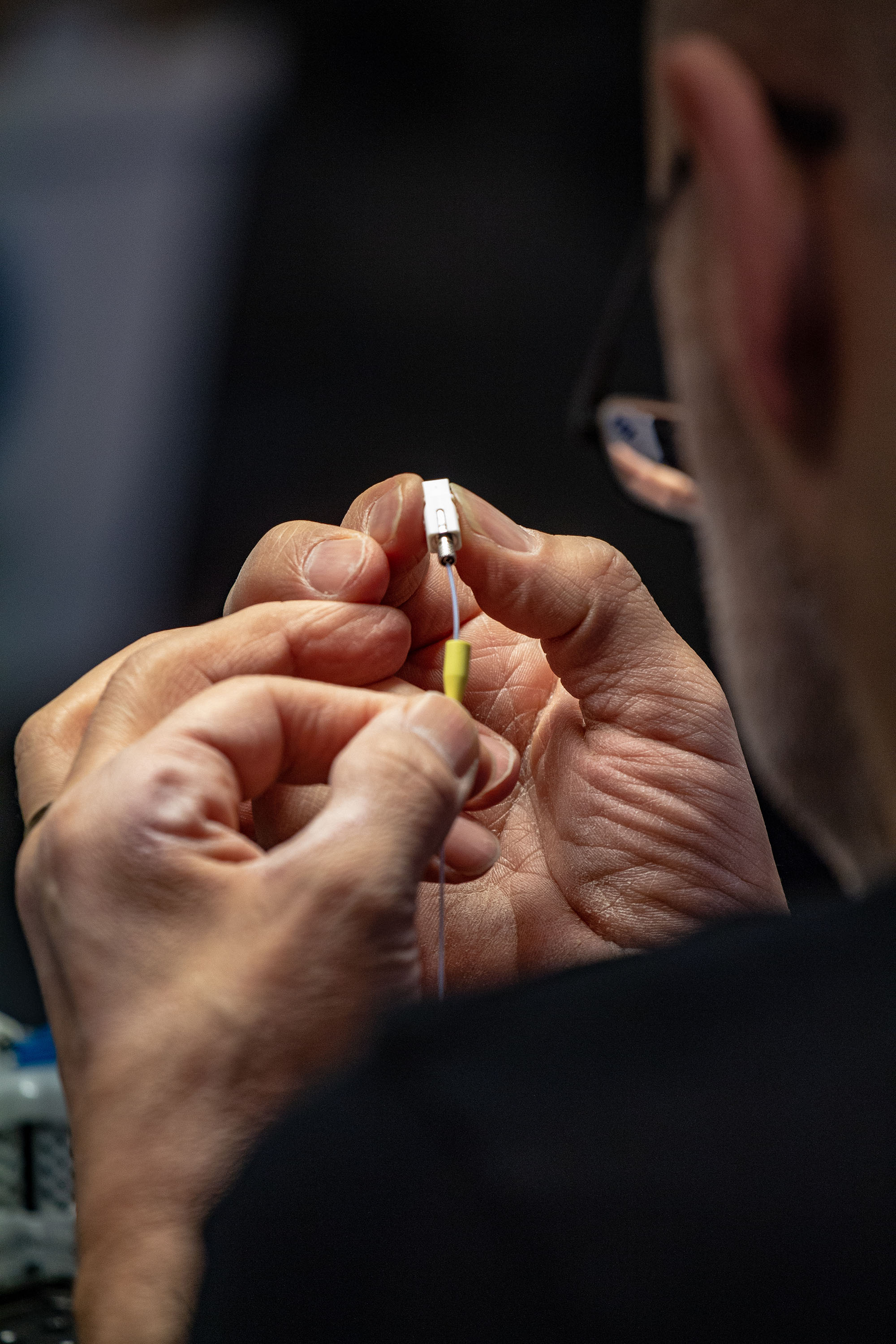 A person is carefully connecting a small, white connector to a fiber optic wire, focusing intently on the task.