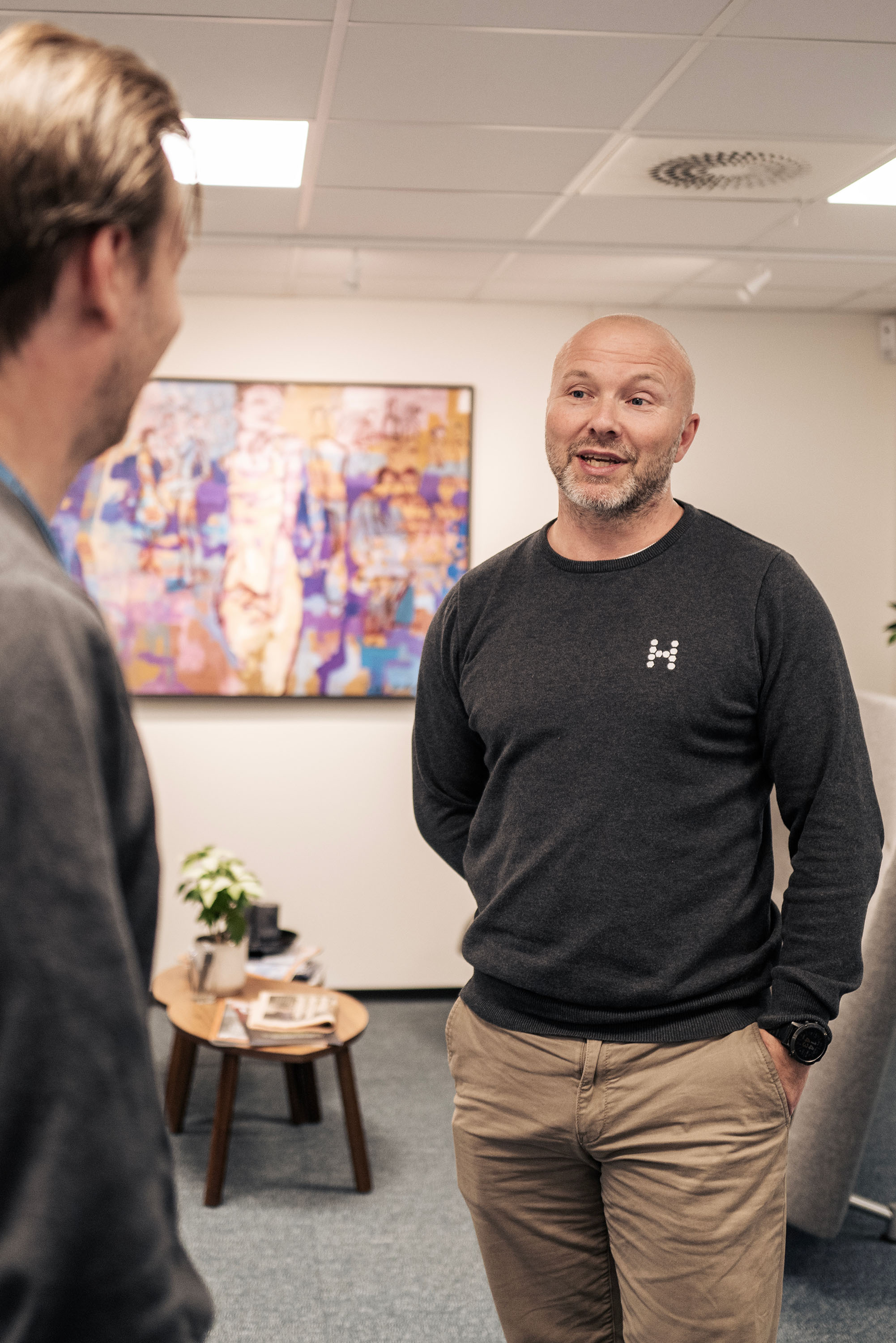 Two people in an office, one in a dark sweater and beige pants. Artwork and a table with a plant are behind them.