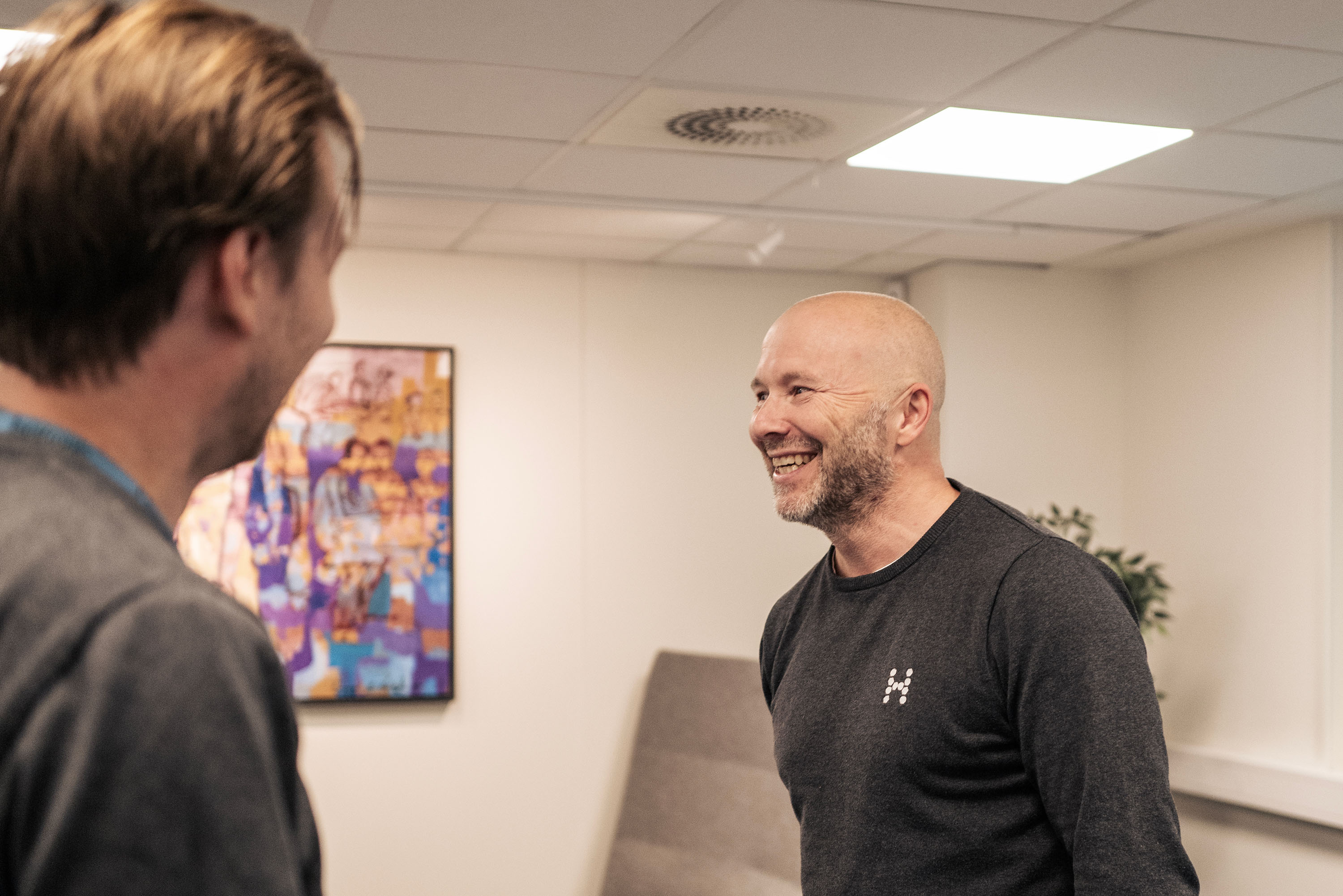 Two people in an office interacts. Artwork and a table with a plant are behind them.