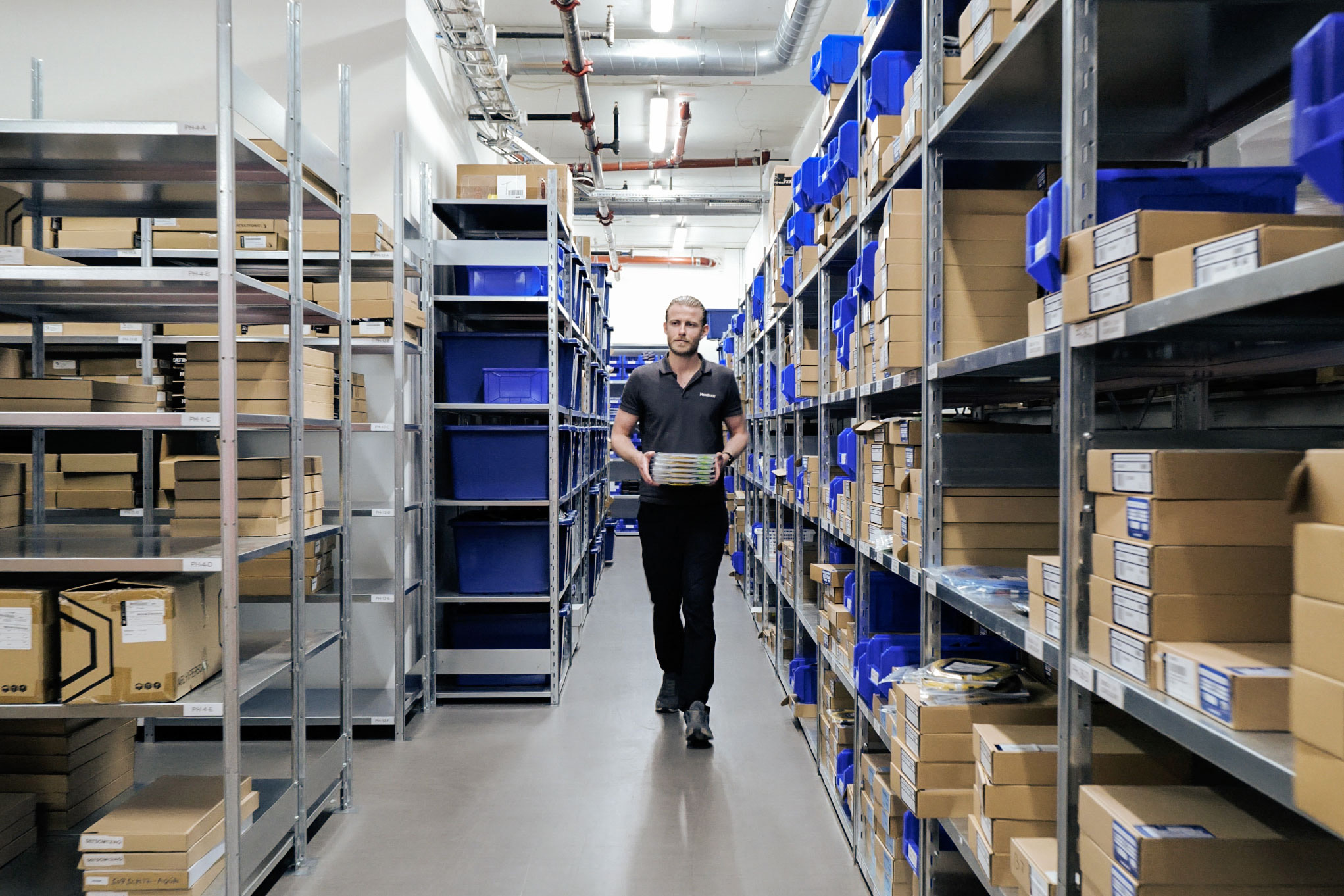 A person in a warehouse aisle, holding boxes, surrounded by shelves filled with organized boxes and bins.
