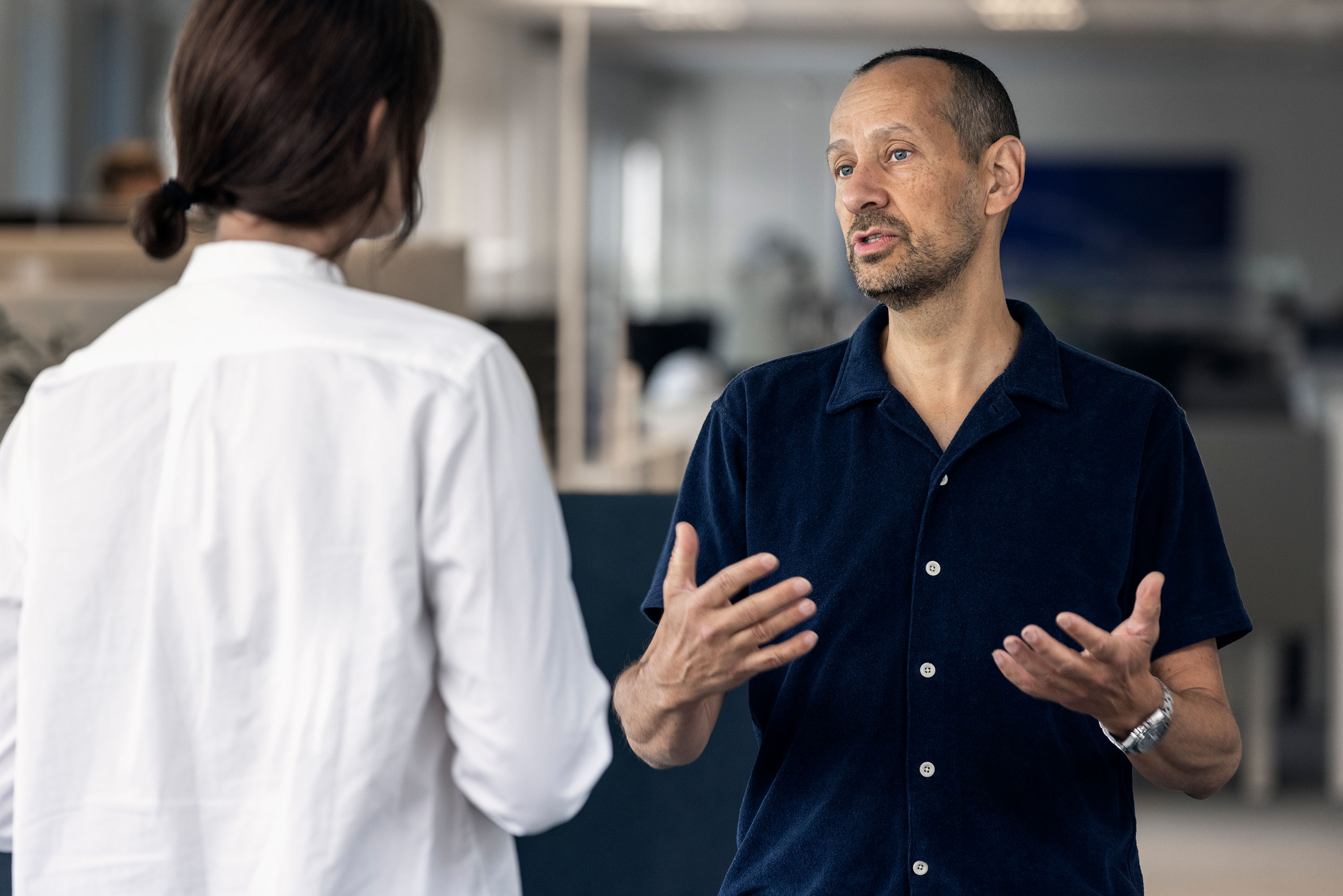 Two people in an office, one facing away in a white shirt, the other in a dark shirt facing the camera, gesturing with hands.