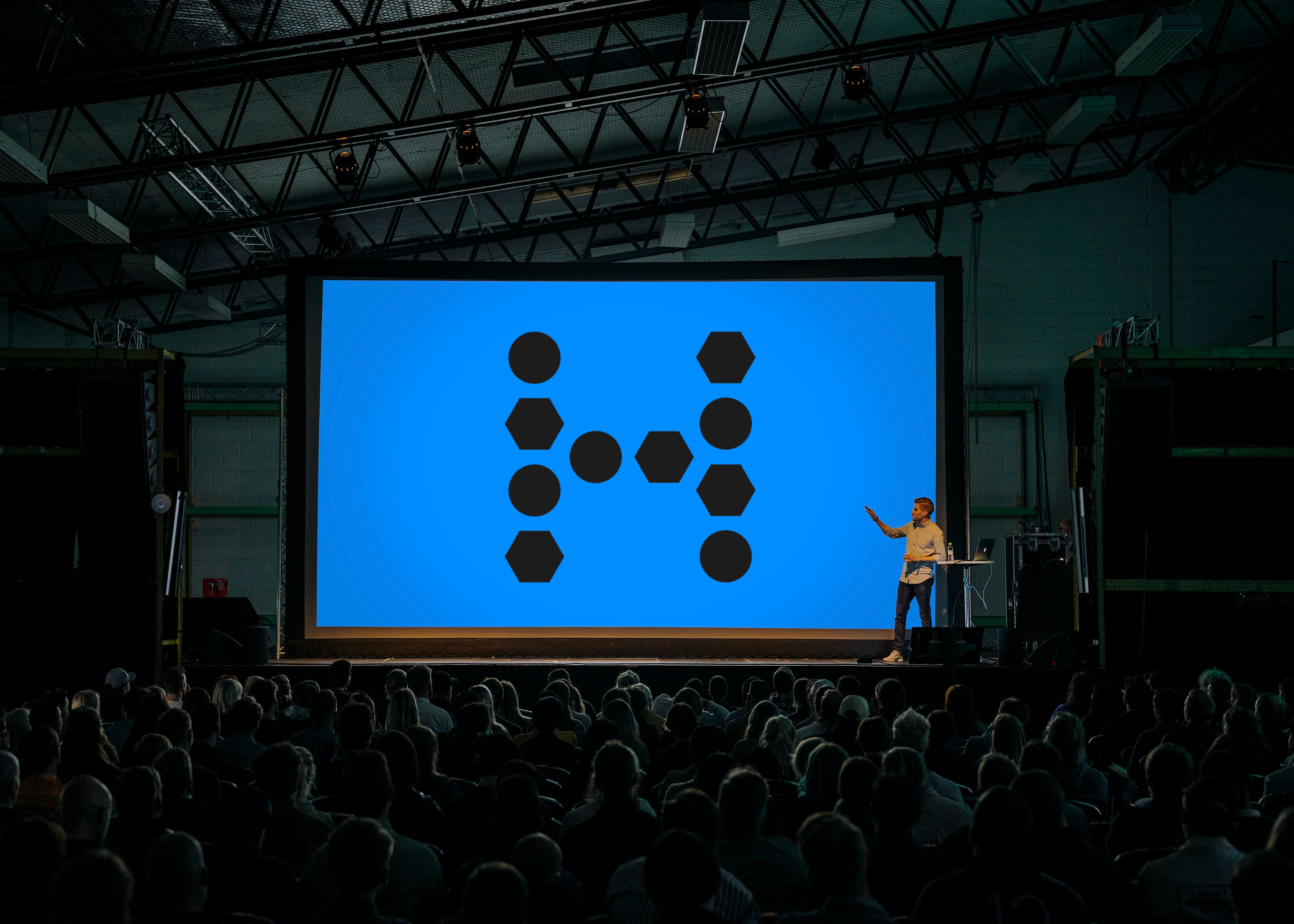 A presenter on a stage points to a large screen with the Hexatronics symbol in black against a blue background. In the foreground a seated audience.
