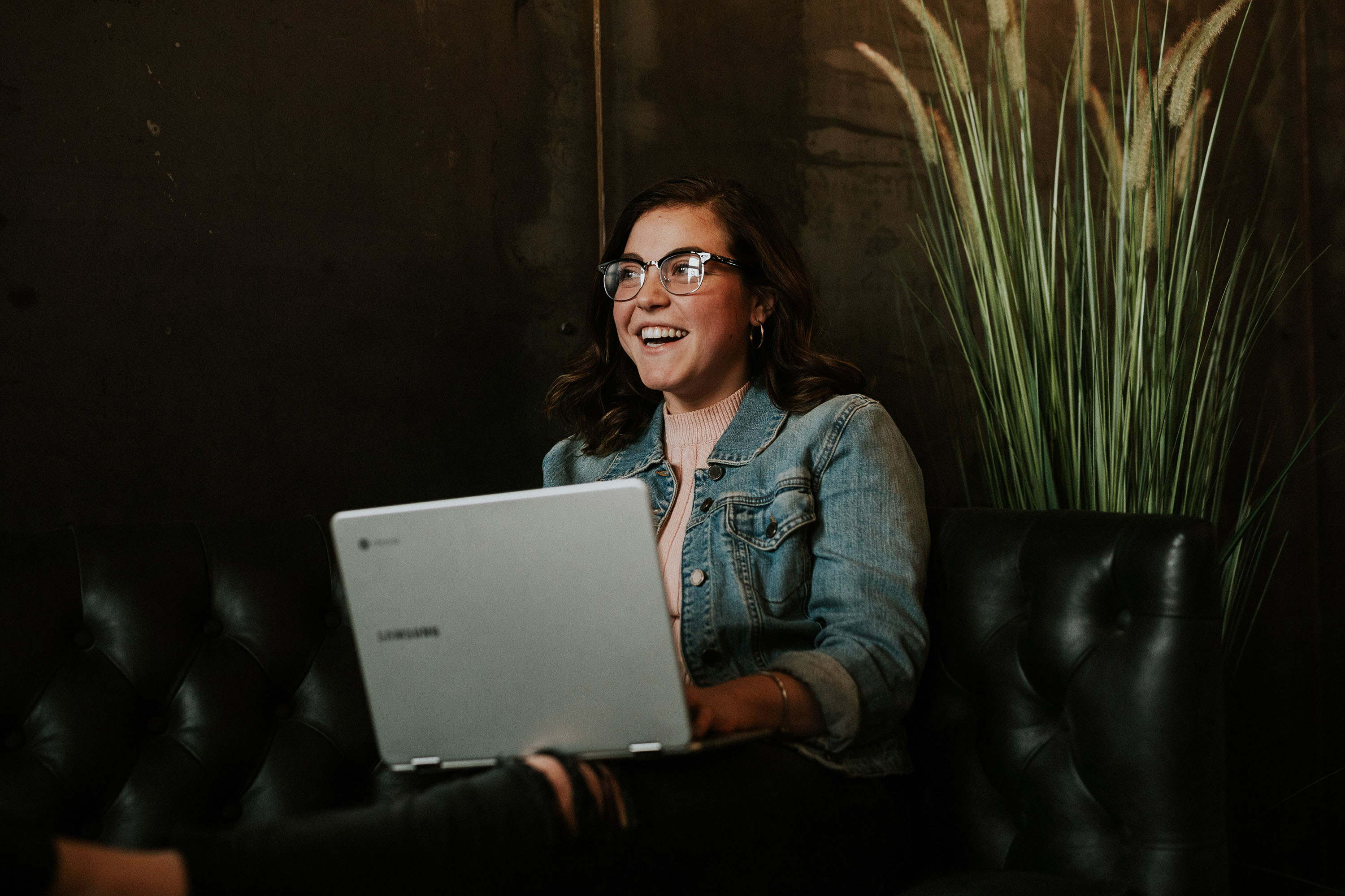 Person in denim jacket on a black couch, using a laptop in a dimly lit room with a tall plant nearby.