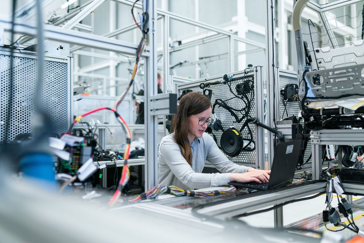 A person is working on a laptop amidst complex machinery and wires, likely in a tech or industrial setting.