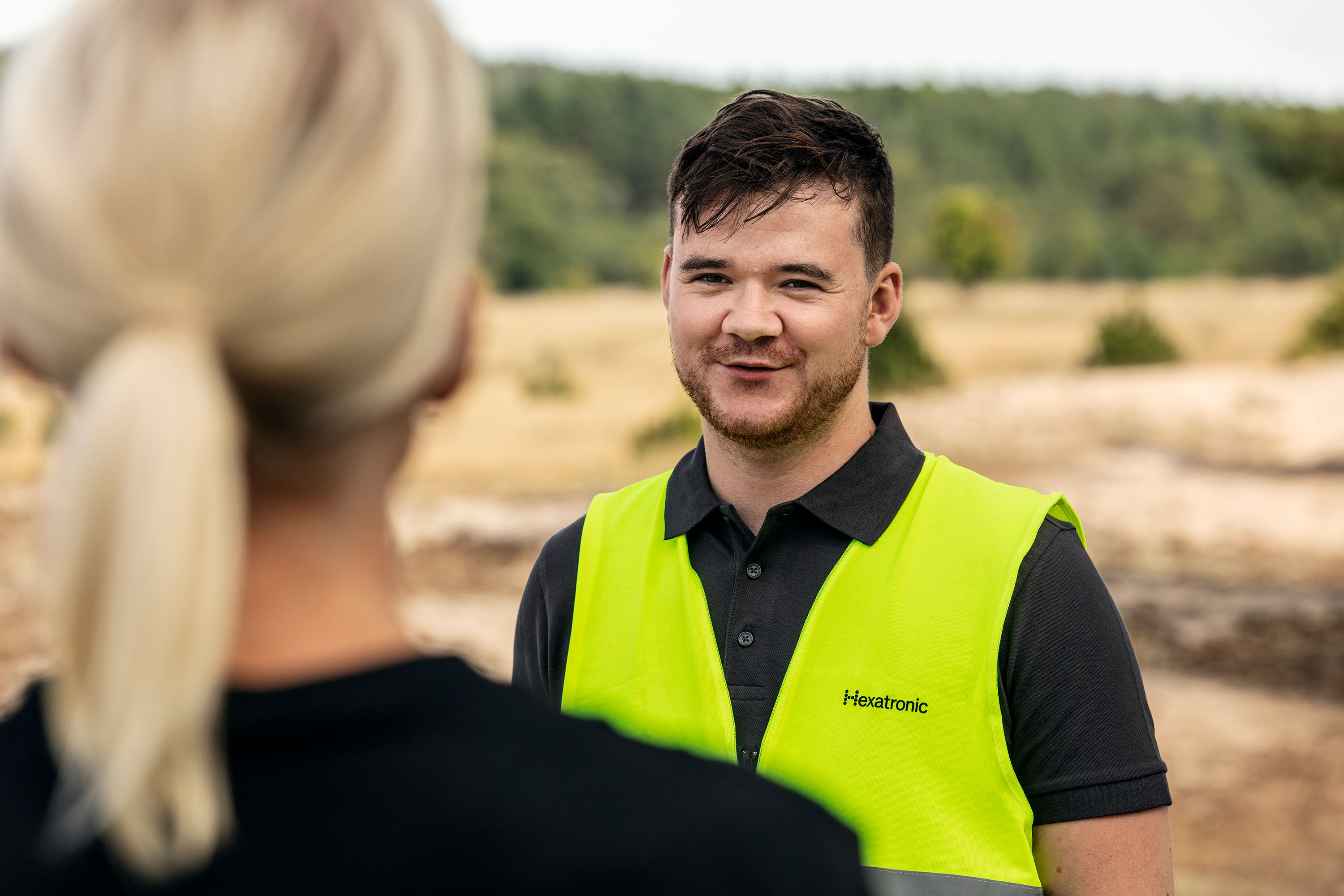 Two people outdoors. One person is wearing a bright yellow safety vest with “Hexatronic” written on it. They appear to be conversing.