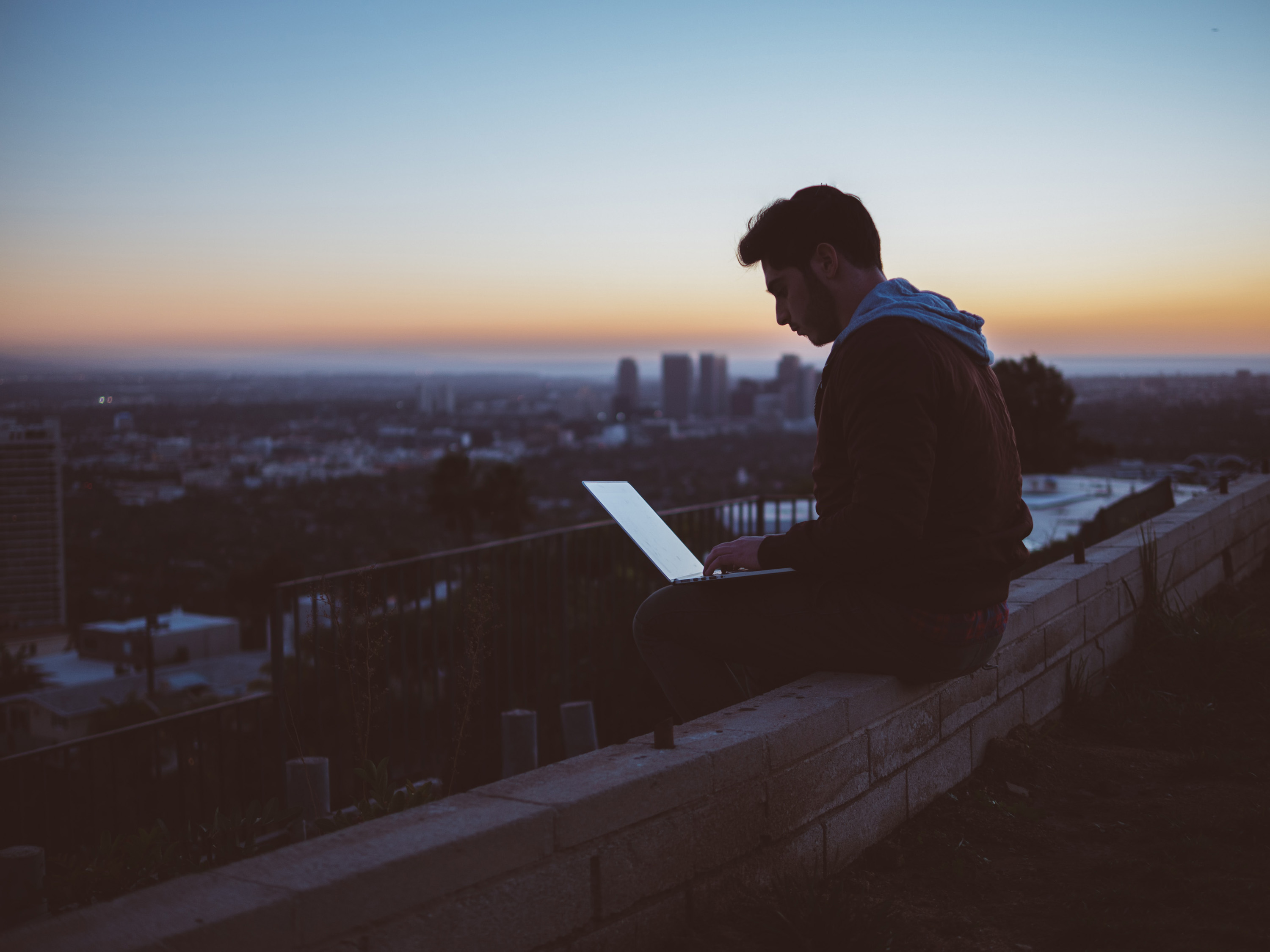 A person is silhouetted against a sunset, sitting on a ledge, working on a laptop with the cityscape in the background.