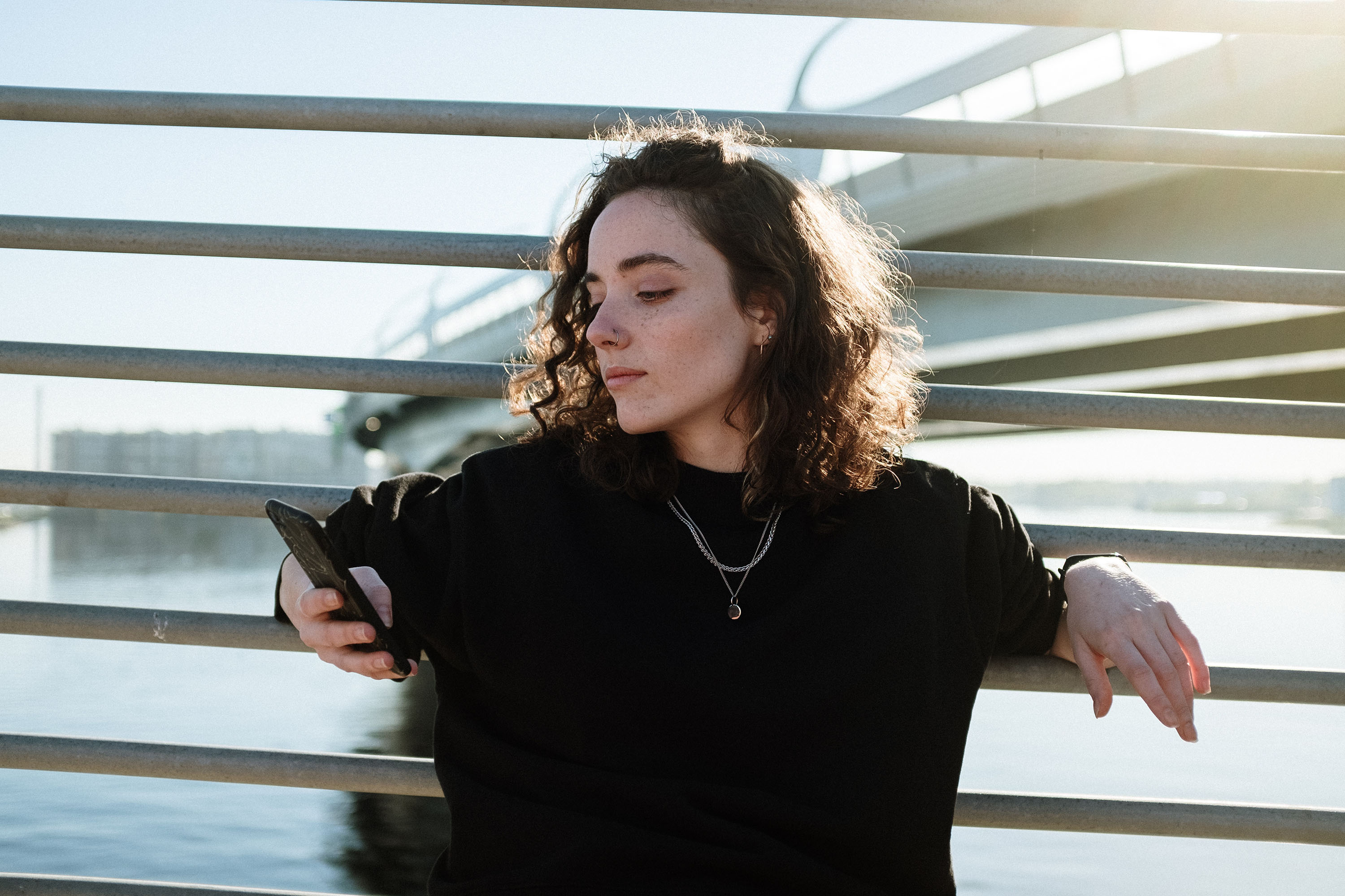 Person leaning on a railing by water, holding a phone, bathed in sunlight. Bridge structure in the background.