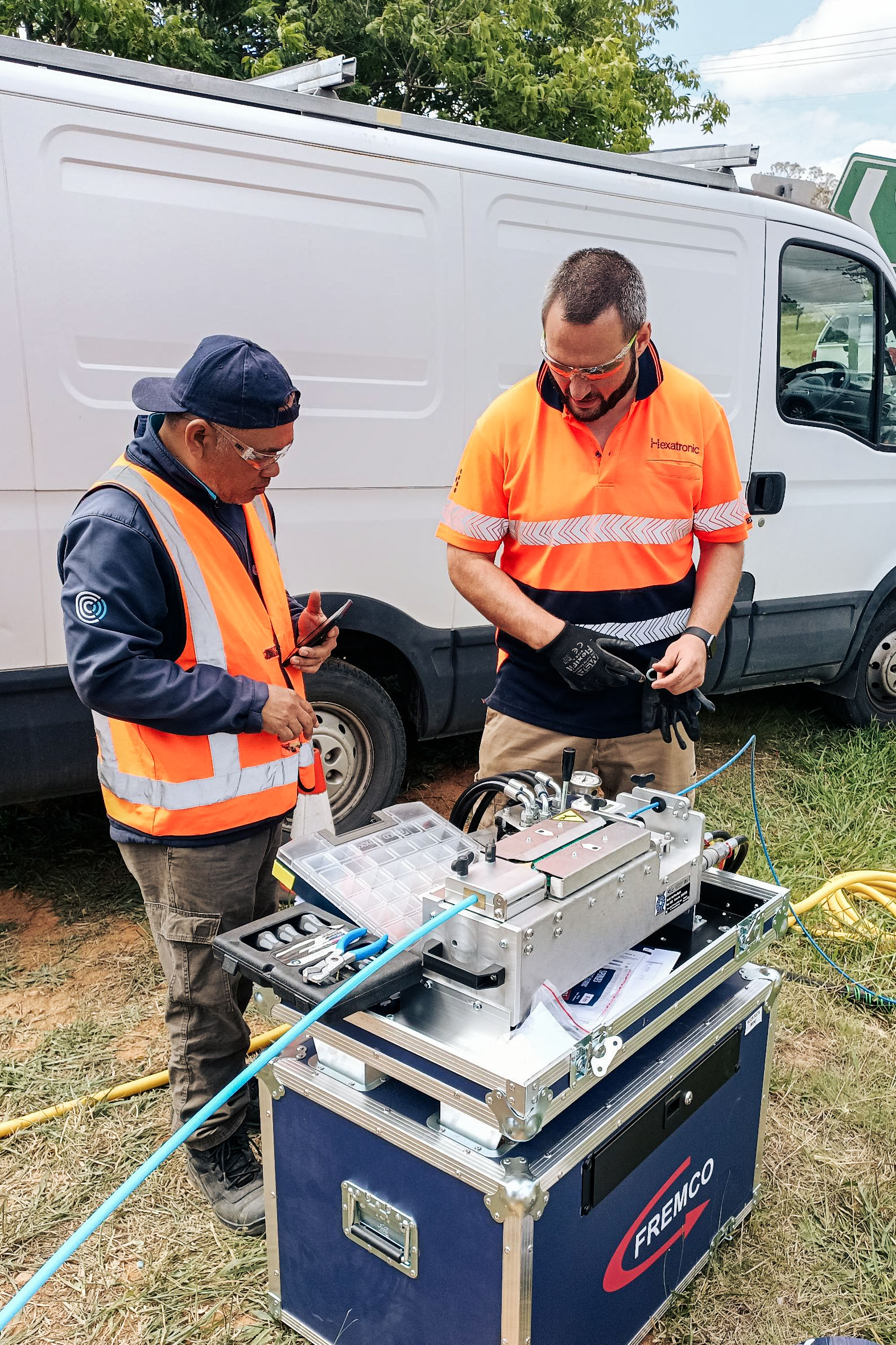 Two workers in safety vests inspecting technical equipment labeled ‘FREMCO’ near a white van, outdoors with greenery in the background
