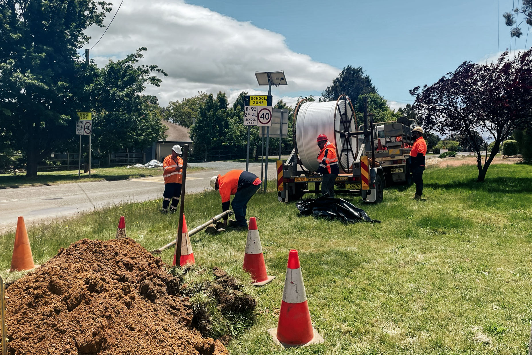 Workers in high-vis gear are digging by a road near traffic cones. A truck and 40 km/h speed limit signs are visible.
