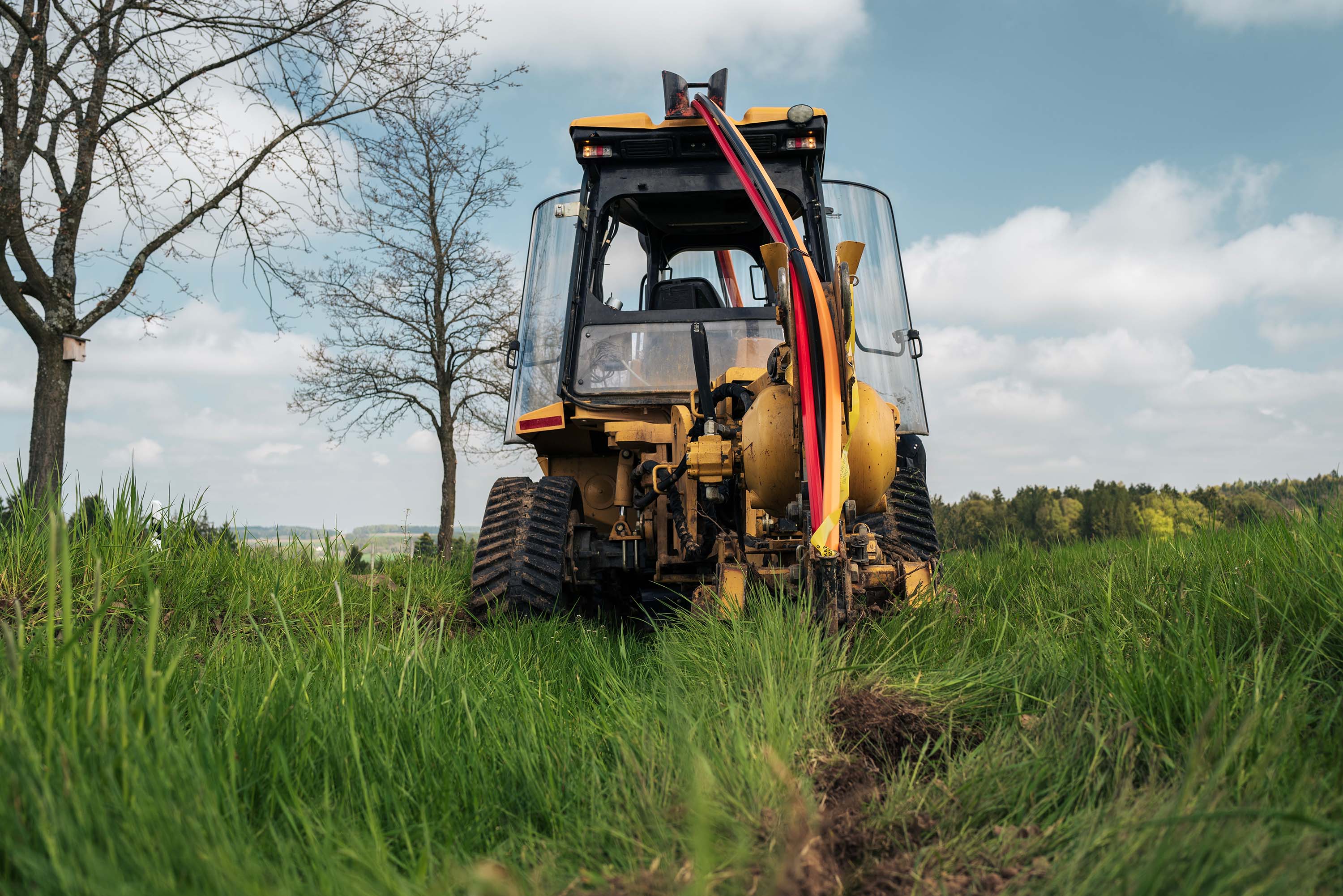 A yellow-black tractor in a lush green field creates a ditch and lays duct, under a clear sky with distant trees.