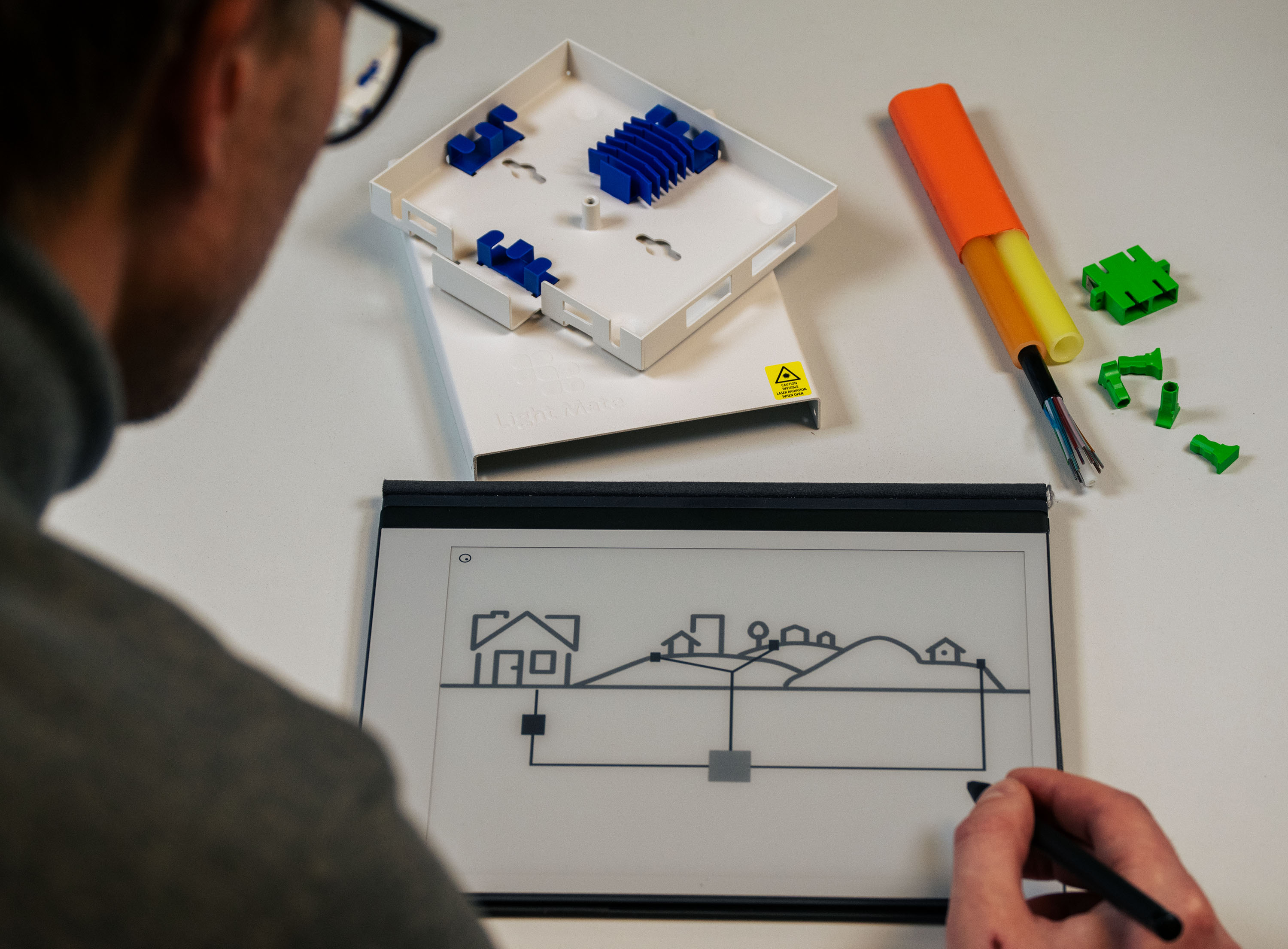 A person drawing a diagram of a fiber network build. Nearby, you can see samples of cables and ducts as well as a wall box.
