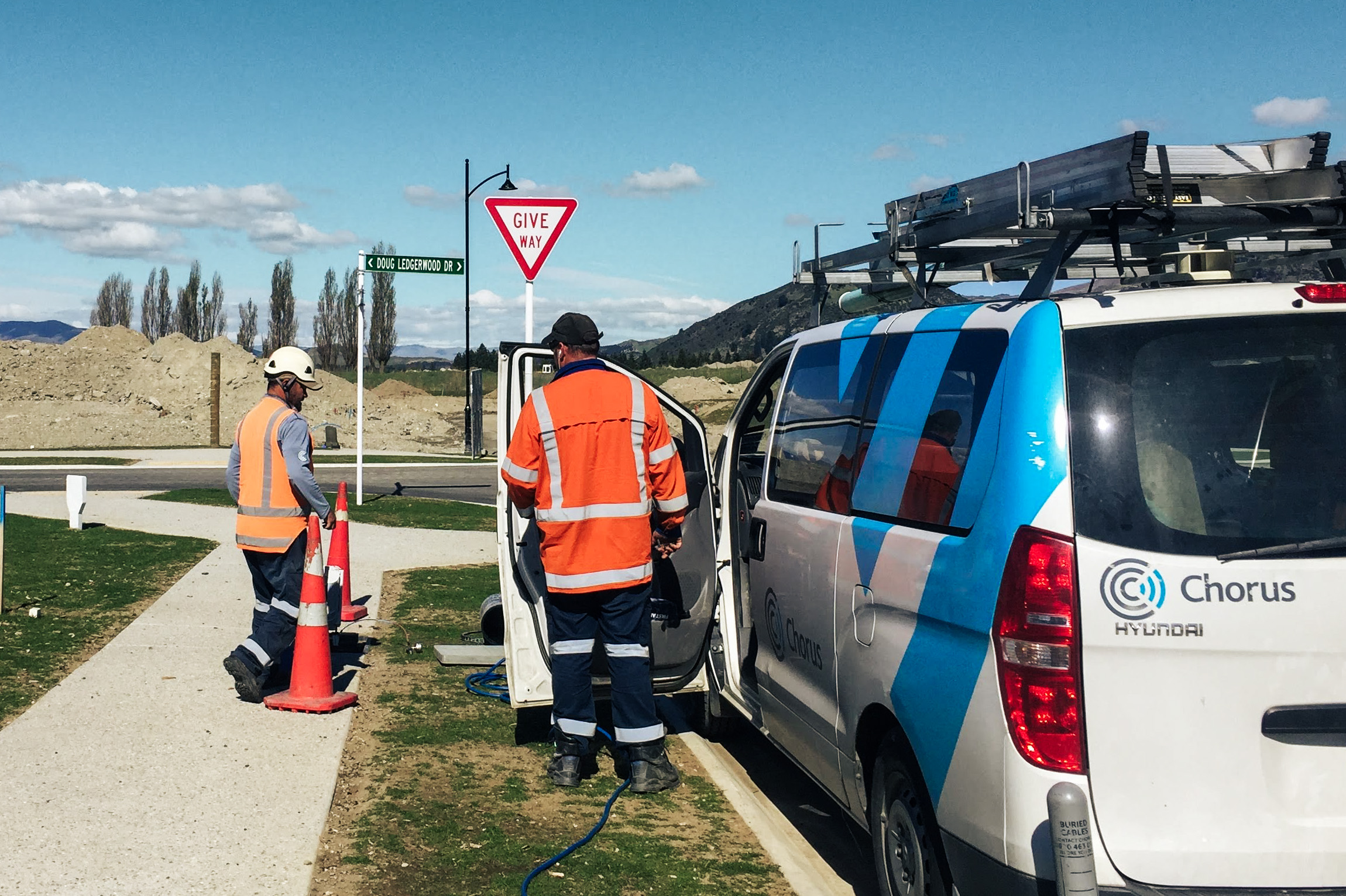Two people in hazard clothing are working on a fiber installation along a road. On the right is a van with the Chorus logo.