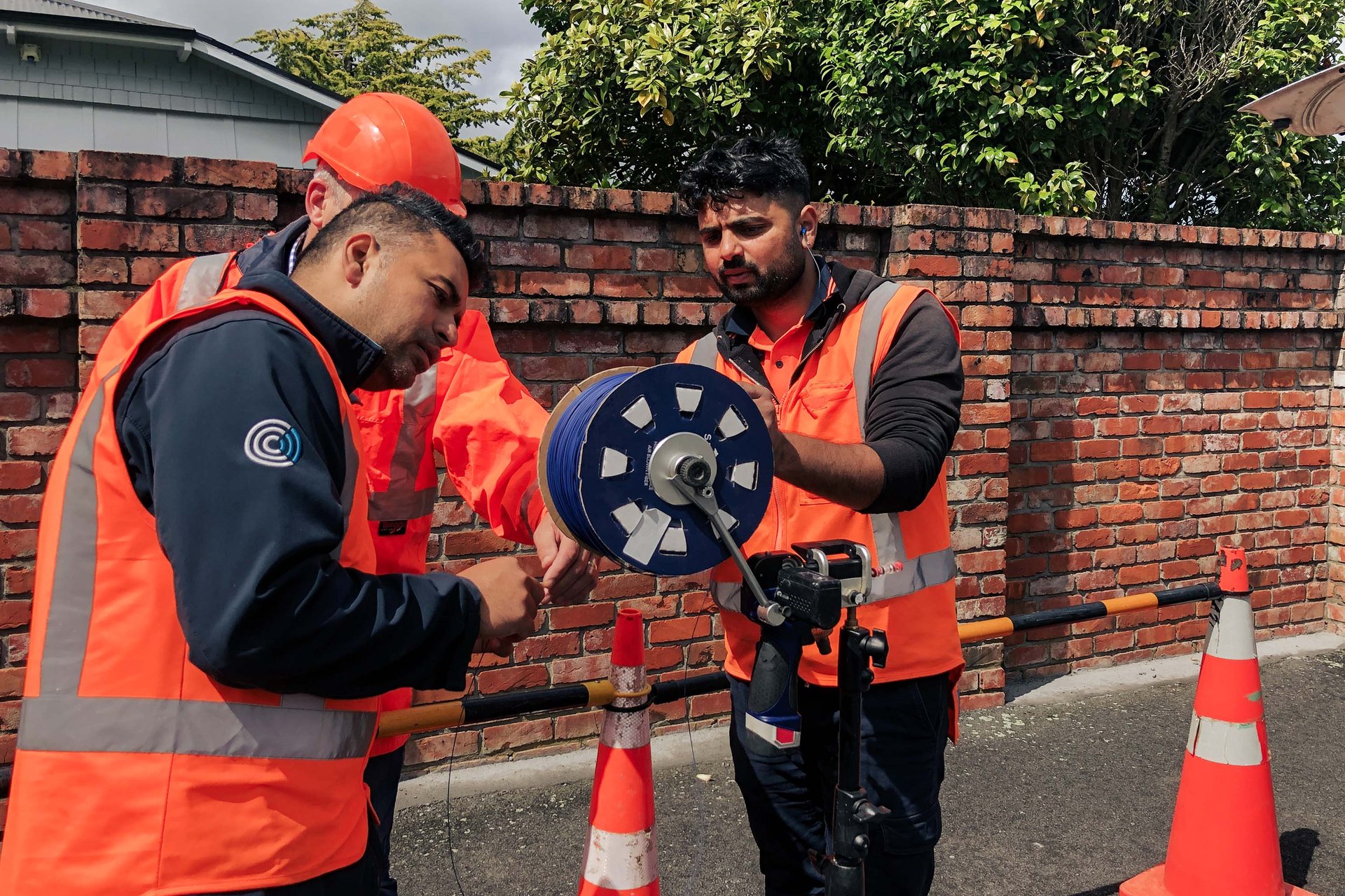 Three people install Stingray Air Blown Fiber. In the background, a house and green trees can be seen behind a red brick wall.