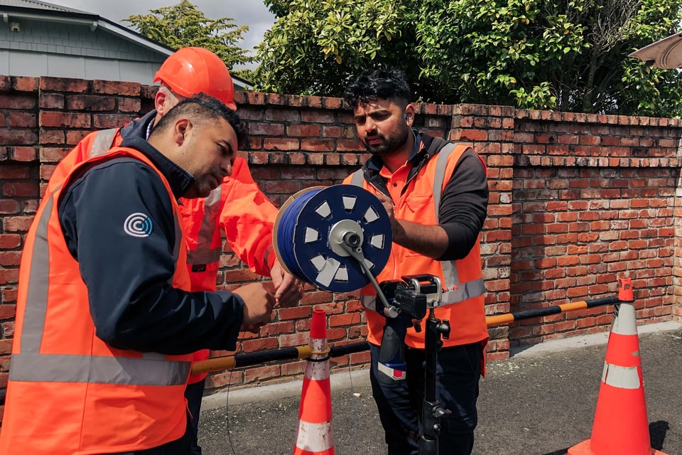 Three people install Stingray Air Blown Fiber. In the background, a house and green trees can be seen behind a red brick wall.