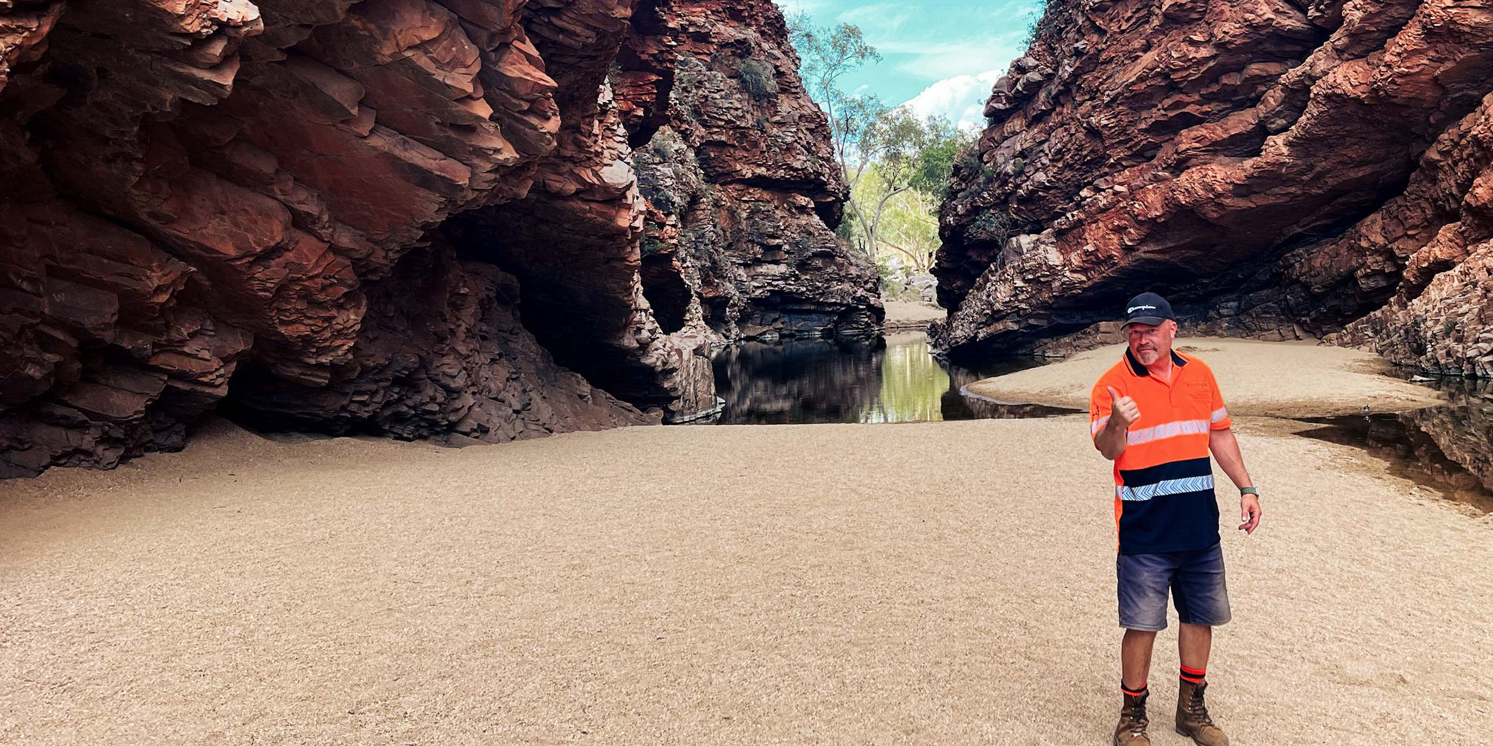 A person in orange work clothes stands on a beach between two rocks and gives the thumbs up.
