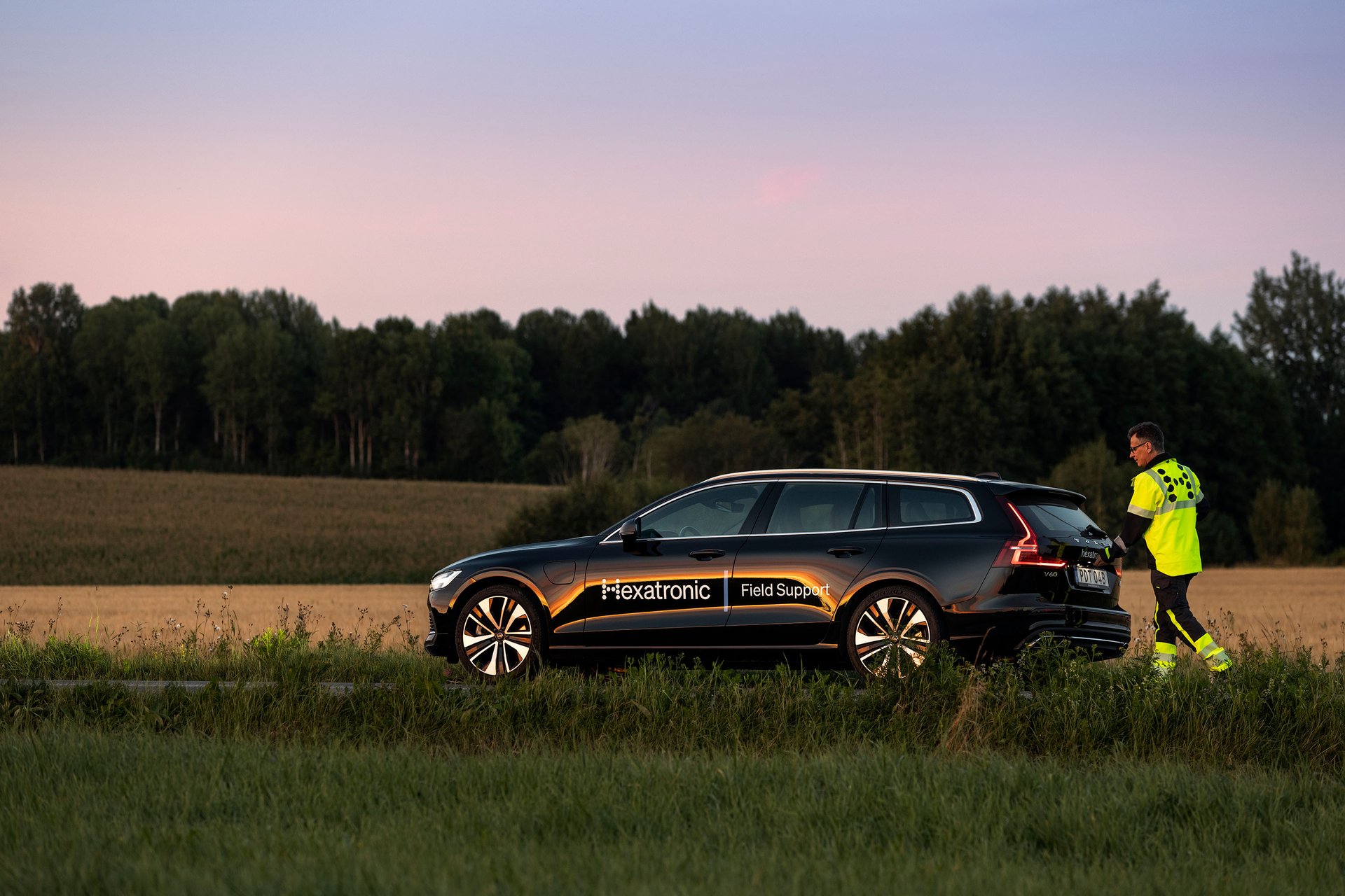 A person in high-vis gear stands next to a ‘Hexatronic Field Support’ car in an open field at dusk.