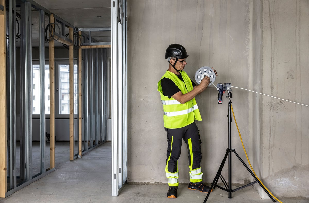 A person in safety gear is installing fiber into a white duct in an unfinished room with exposed framing and wiring.