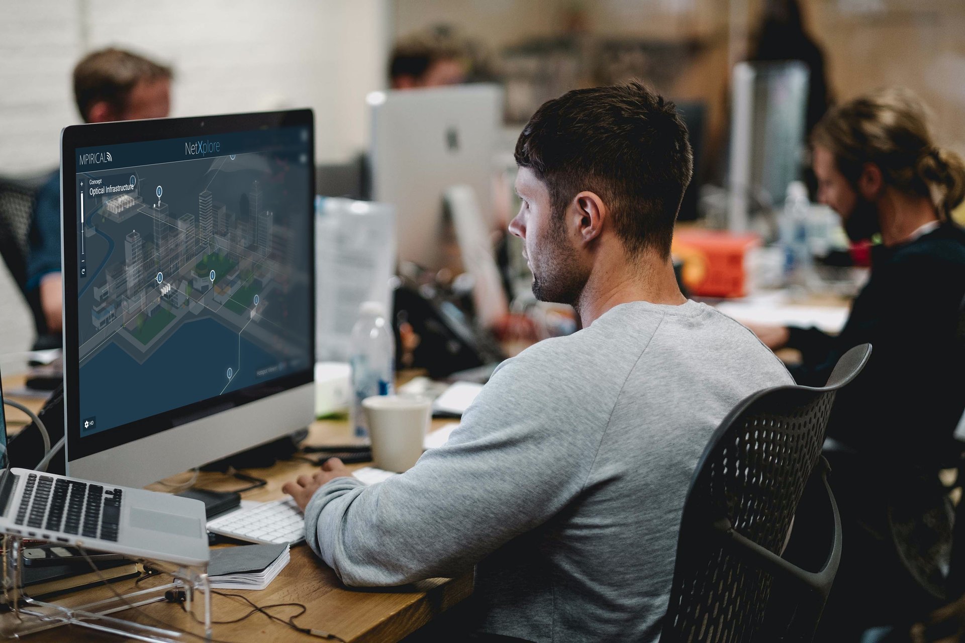 A person is working on a computer displaying network plans in a busy office with colleagues around.