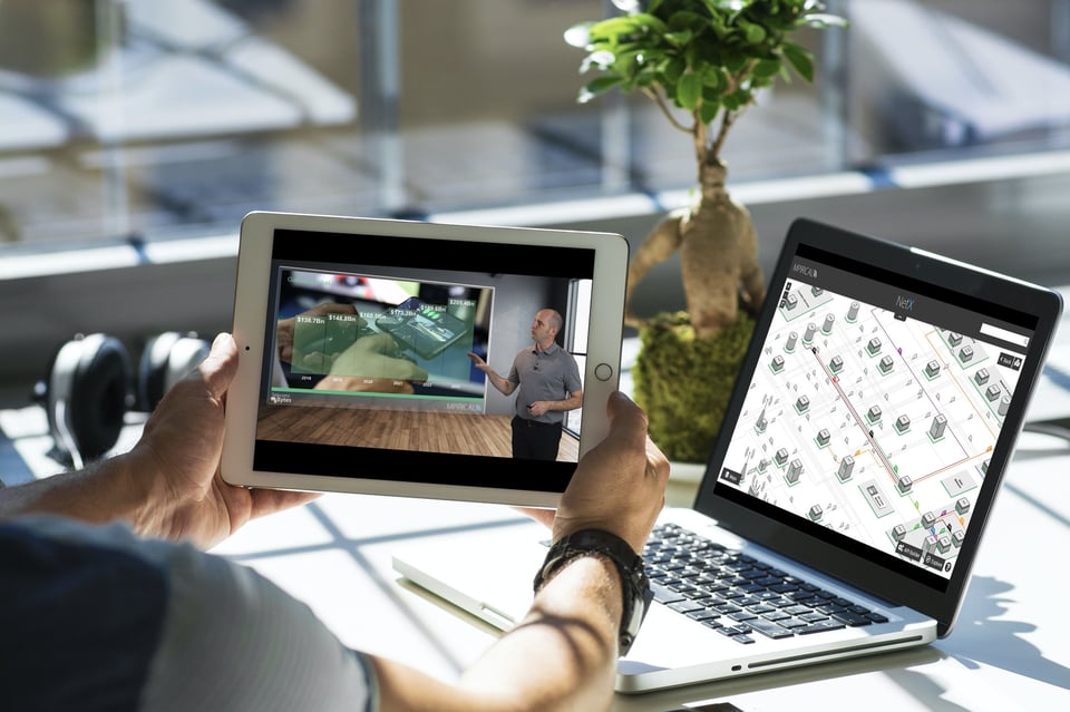 A person multitasks on a tablet showing a video and a laptop with a map showing a wireless network, placed on a table near a window.