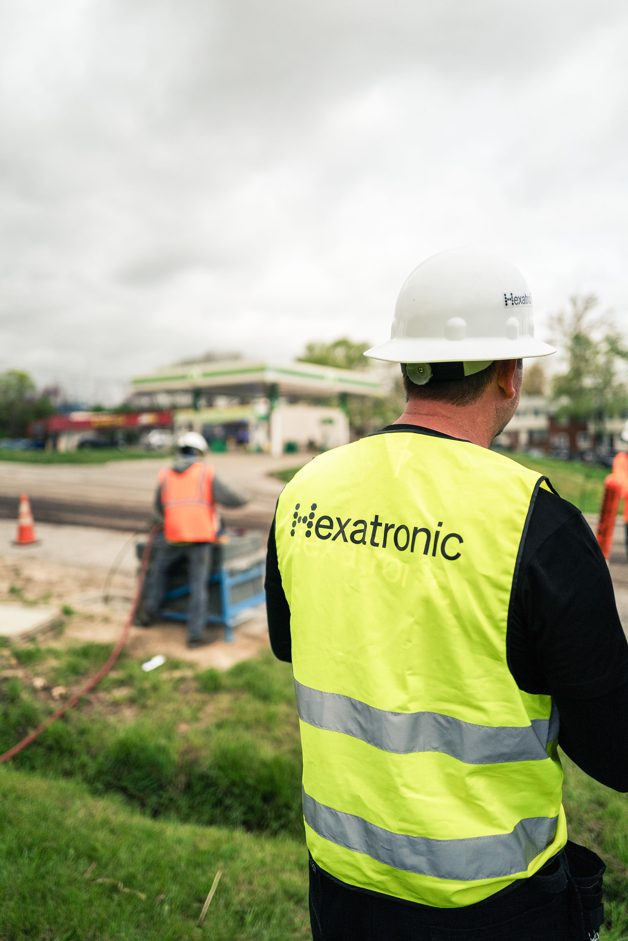 A worker in a Hexatronic vest and helmet at an outdoor construction site under cloudy skies.