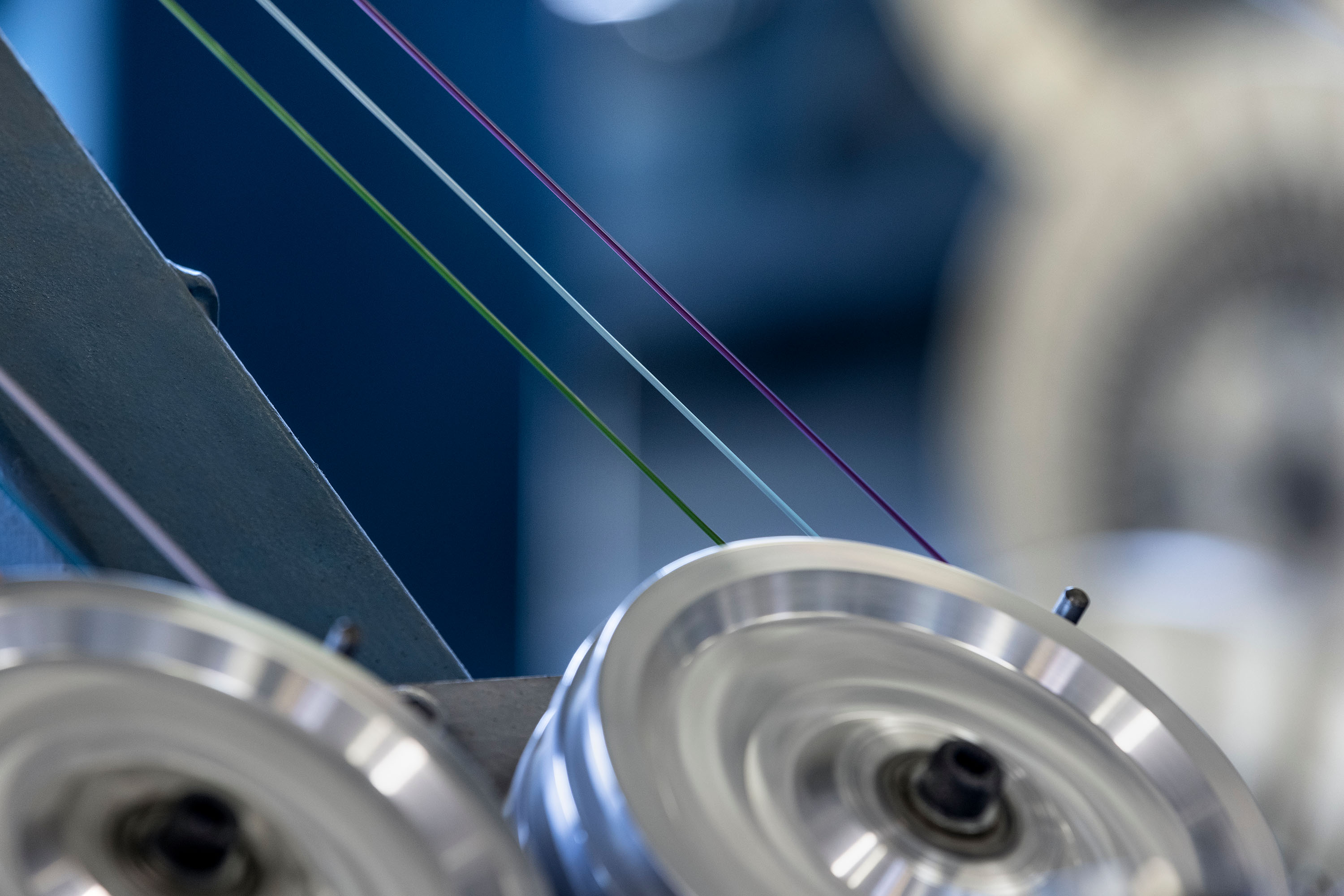 Close-up of industrial machinery with colorful fiber optic threads running over spinning wheels, likely for cable manufacturing.