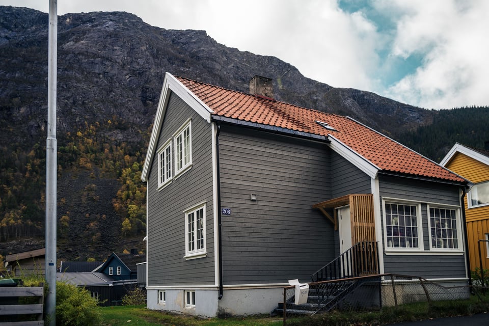 A grey two-story house with a red roof is nestled at the foot of a rocky mountain, surrounded by greenery and similar homes.