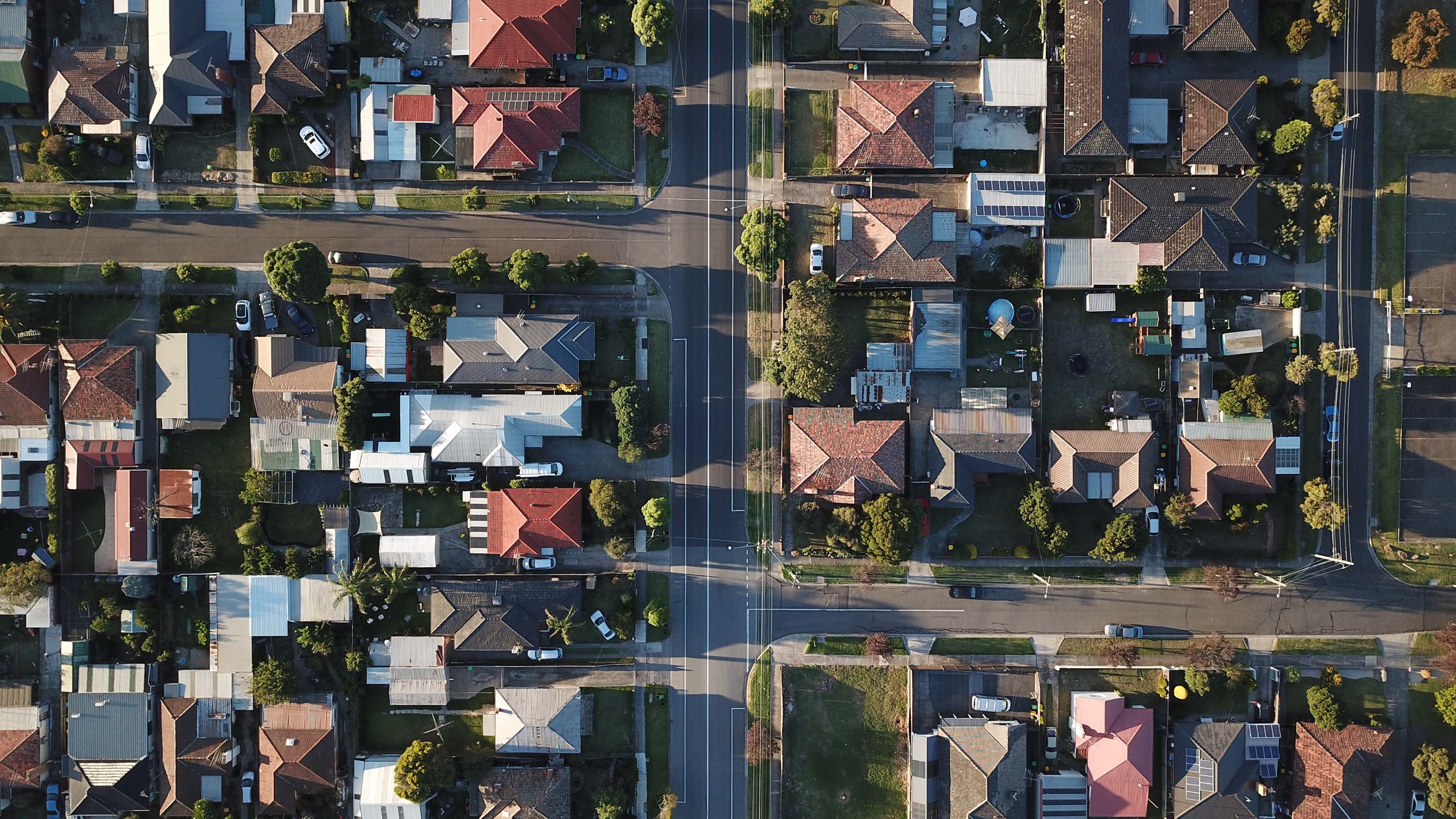 Aerial view of a suburban neighborhood, various houses, green lawns, intersecting roads, bathed in soft sunlight.